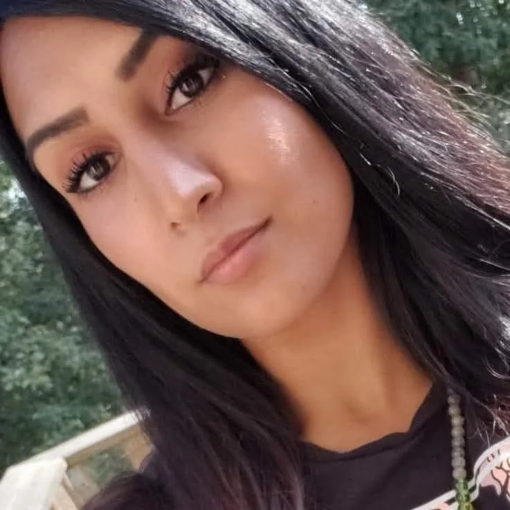 Close-up of a woman with long black hair, wearing a black shirt with a necklace, outdoors with green foliage in the background.