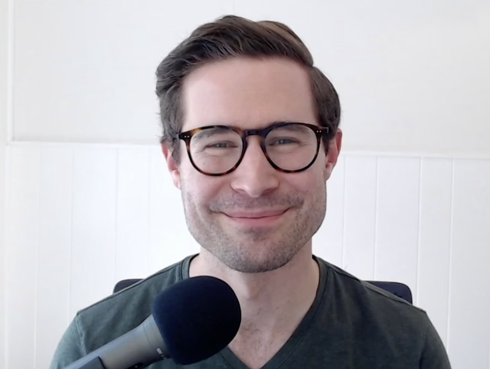A man with brown hair, glasses, and a slight smile, sitting at a desk with a microphone in front of him, in front of a white wall.