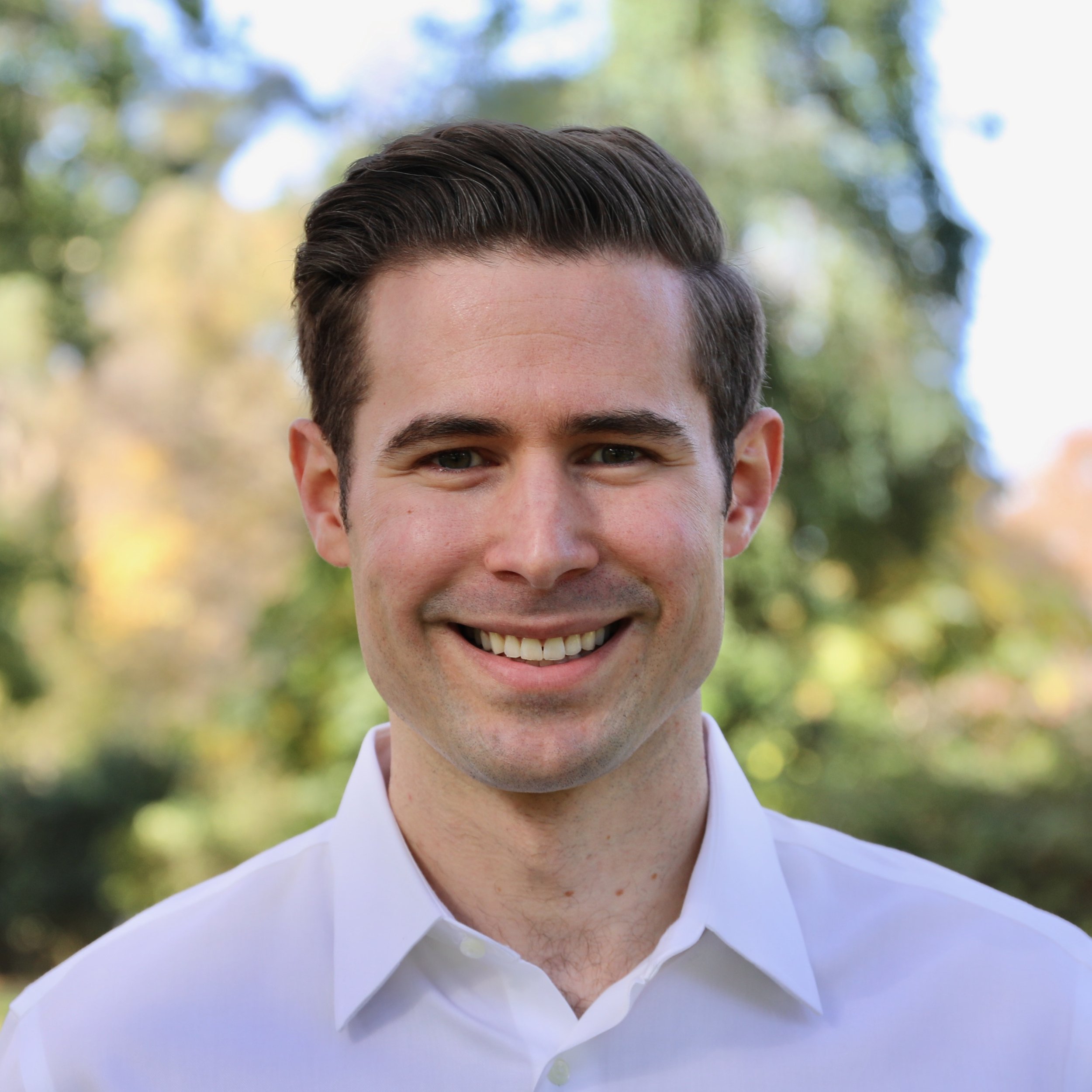 A smiling young man with brown hair and wearing a white shirt, outdoors with blurred trees and sunlight in the background.