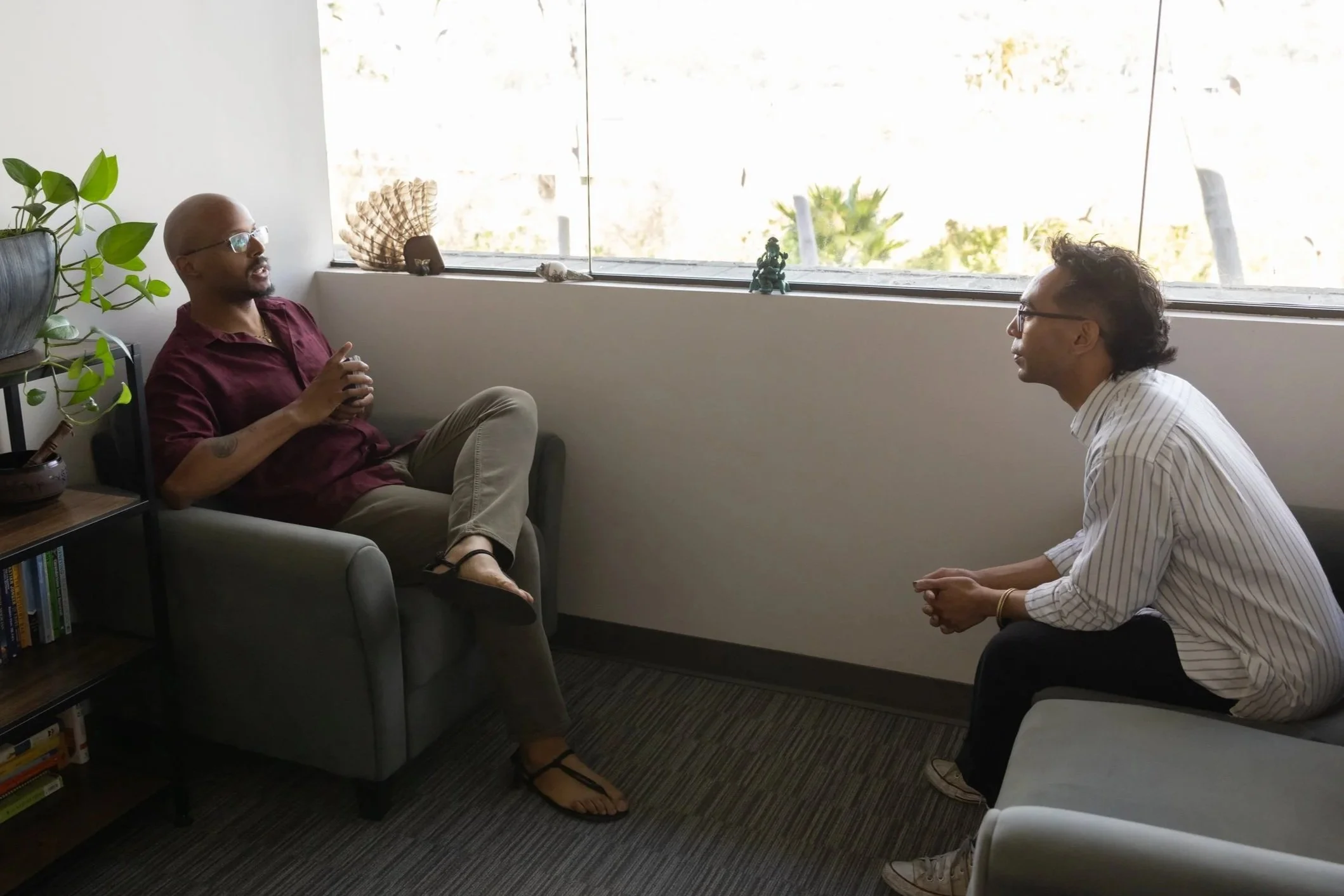 Two men are having a conversation in an office with large windows. One is sitting on a chair, and the other is kneeling on a stool. There are decorative pieces and plants on the window sill.