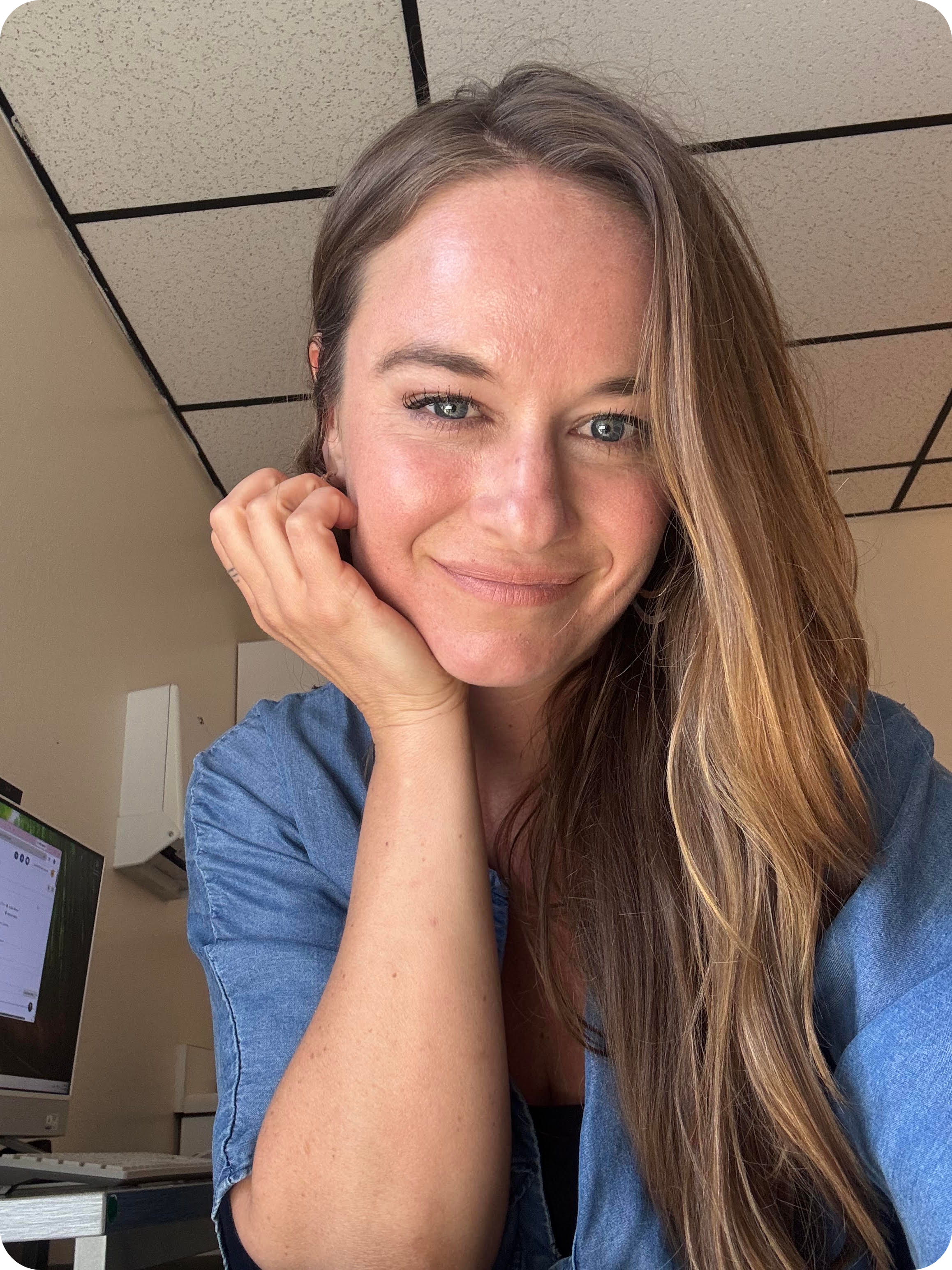 Close-up selfie of a smiling woman with long highlighted brown hair wearing a blue shirt indoors