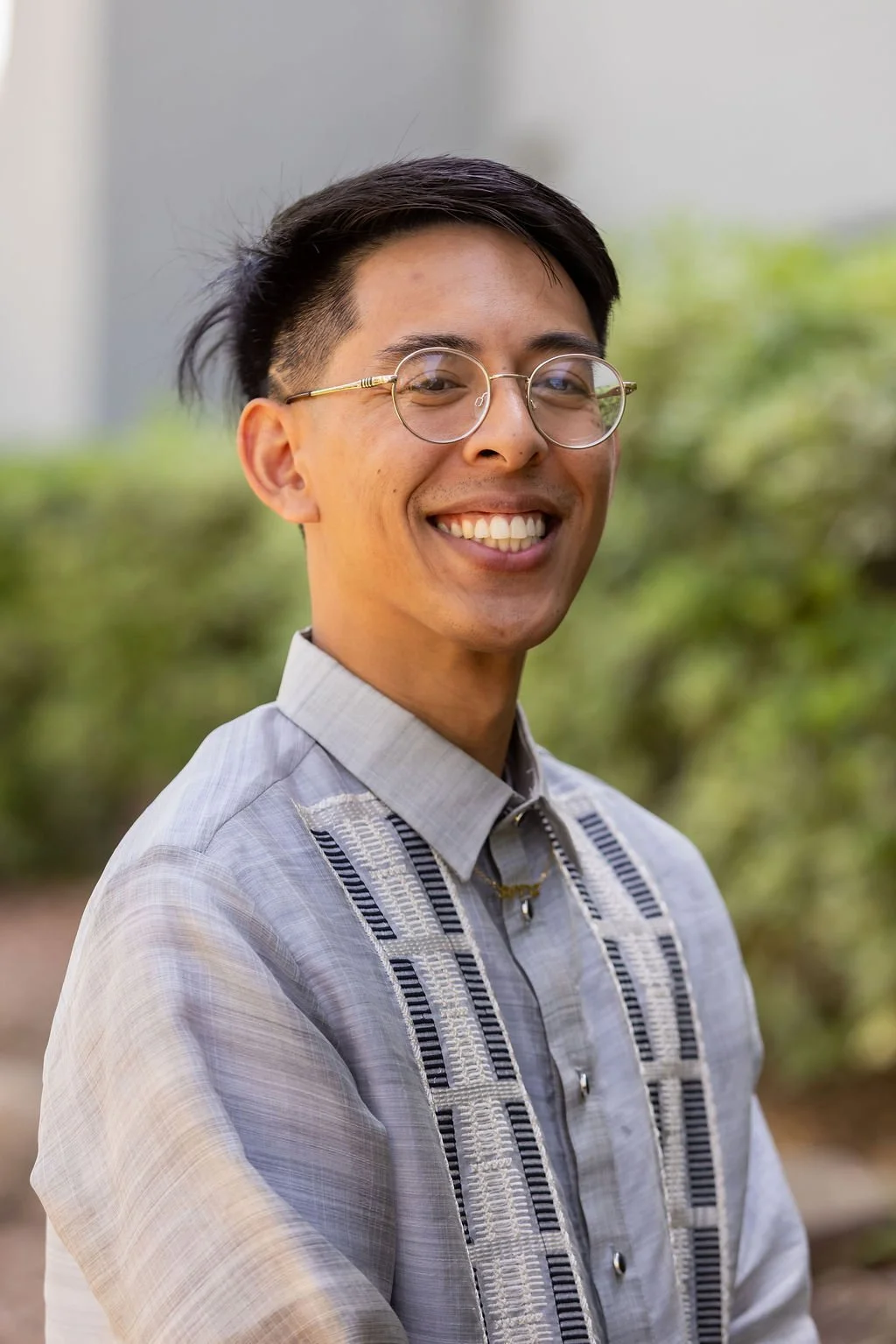 Smiling man wearing round glasses and a light gray embroidered shirt outdoors