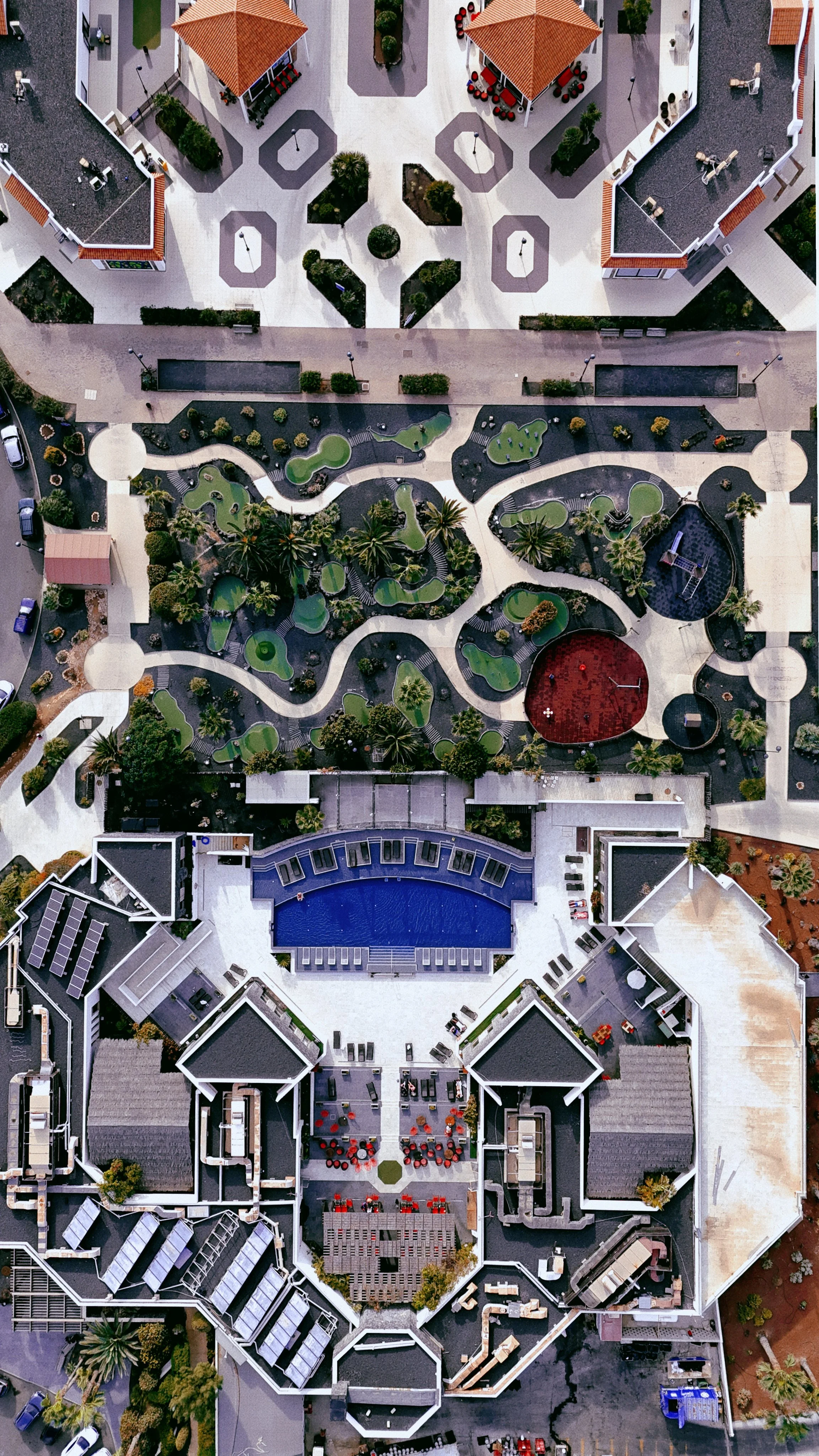Vista aerea di un complesso architettonico con tetti rossi, cortili, giardini, percorsi e una piscina blu centrale.