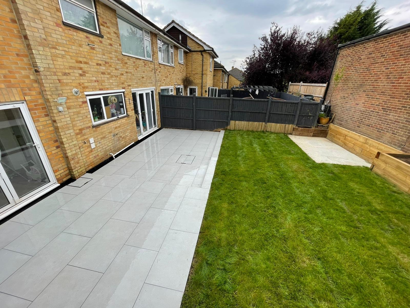 Backyard with a brick house, large patio area with light gray tiles, green lawn, wooden planters, and a black fence.