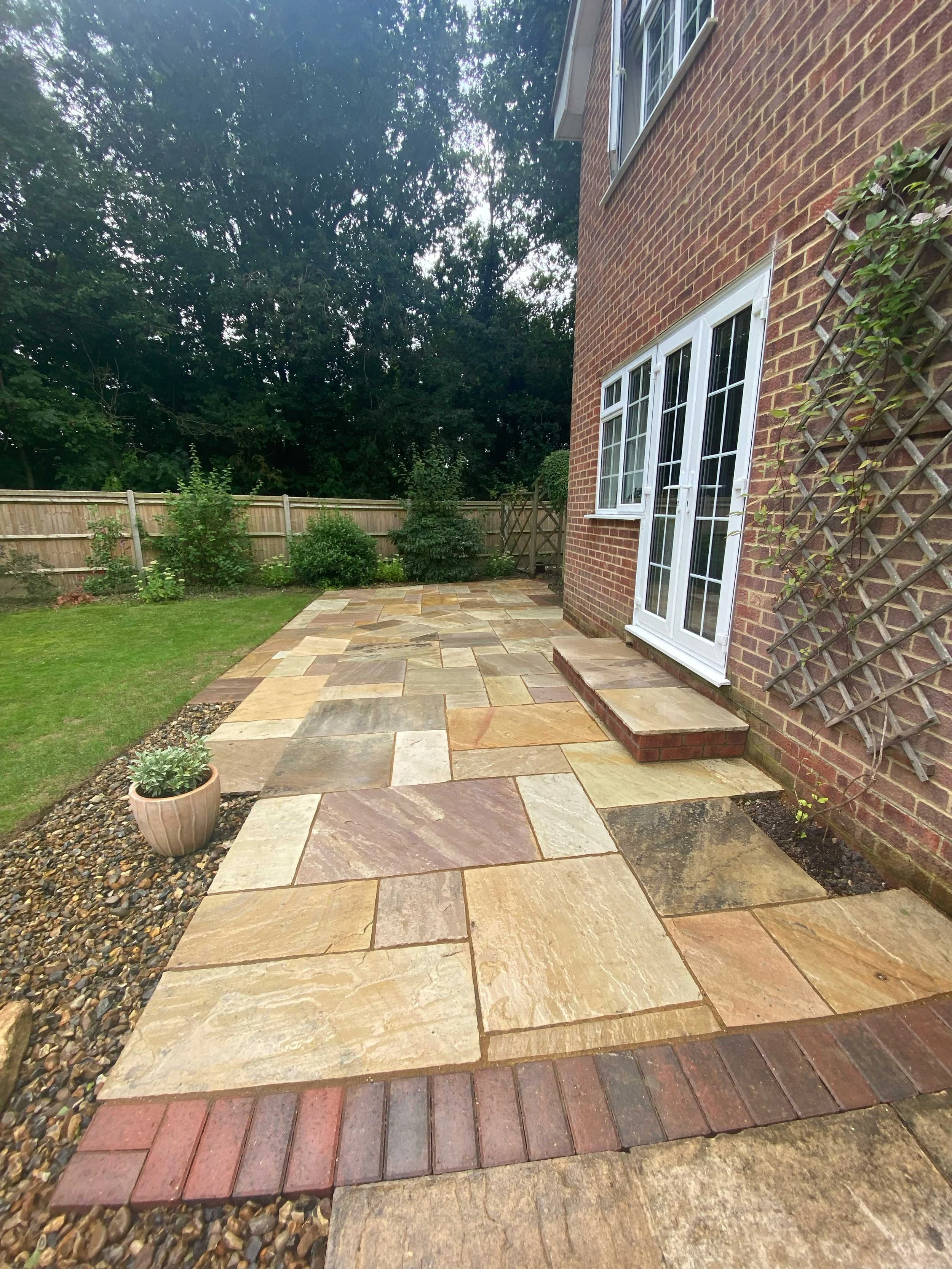 Backyard patio with brick wall and stone paving next to green lawn and wooden fence.