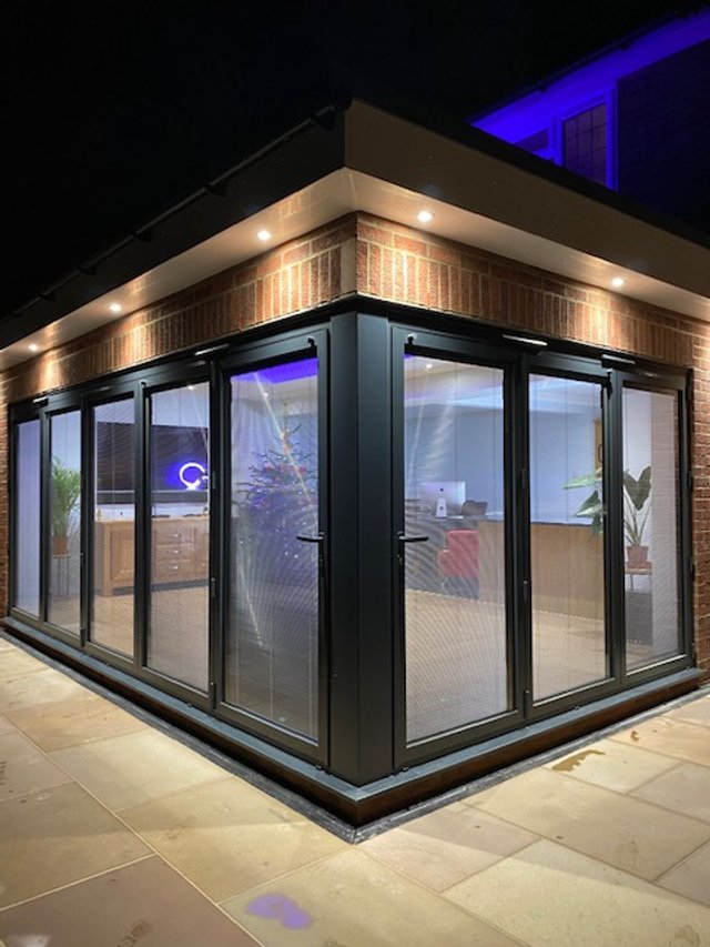 Modern glass-walled room with bi-fold doors and indoor plants, illuminated at night.