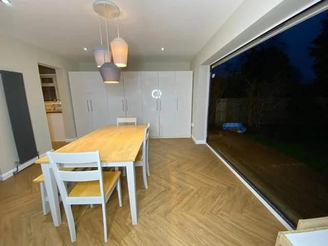 Modern dining room with a wooden table and white chairs, large floor-to-ceiling window overlooking a dark outdoor space, and sleek white cabinets.