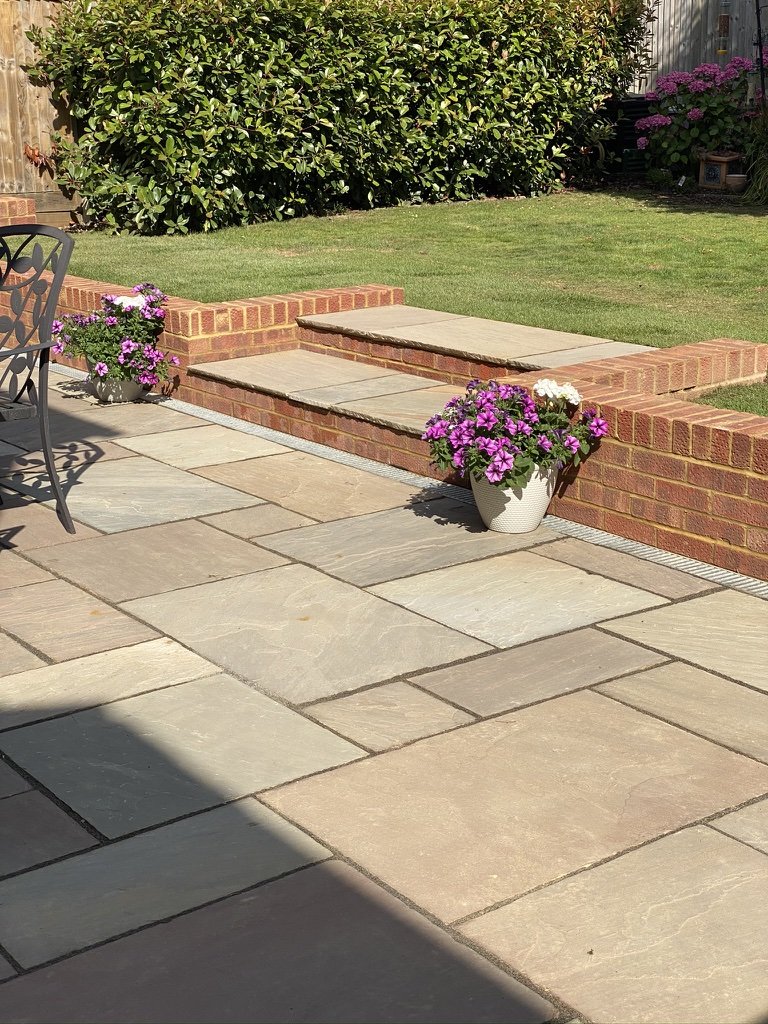 Paved patio with brick steps, potted purple flowers, and a grassy lawn in the background.