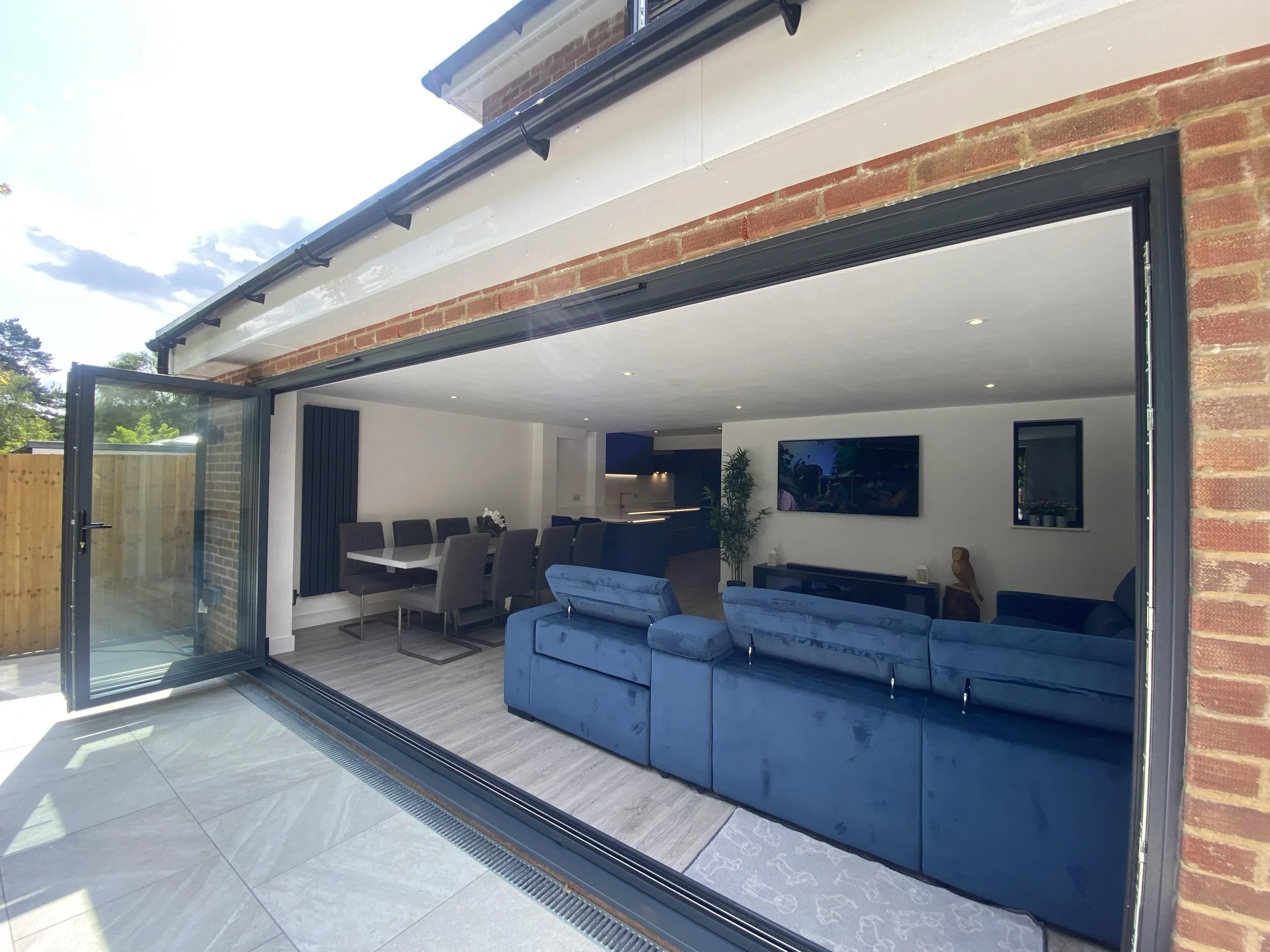 Modern living room with blue sofa, open bi-fold glass doors, dining table, and TV on wall.