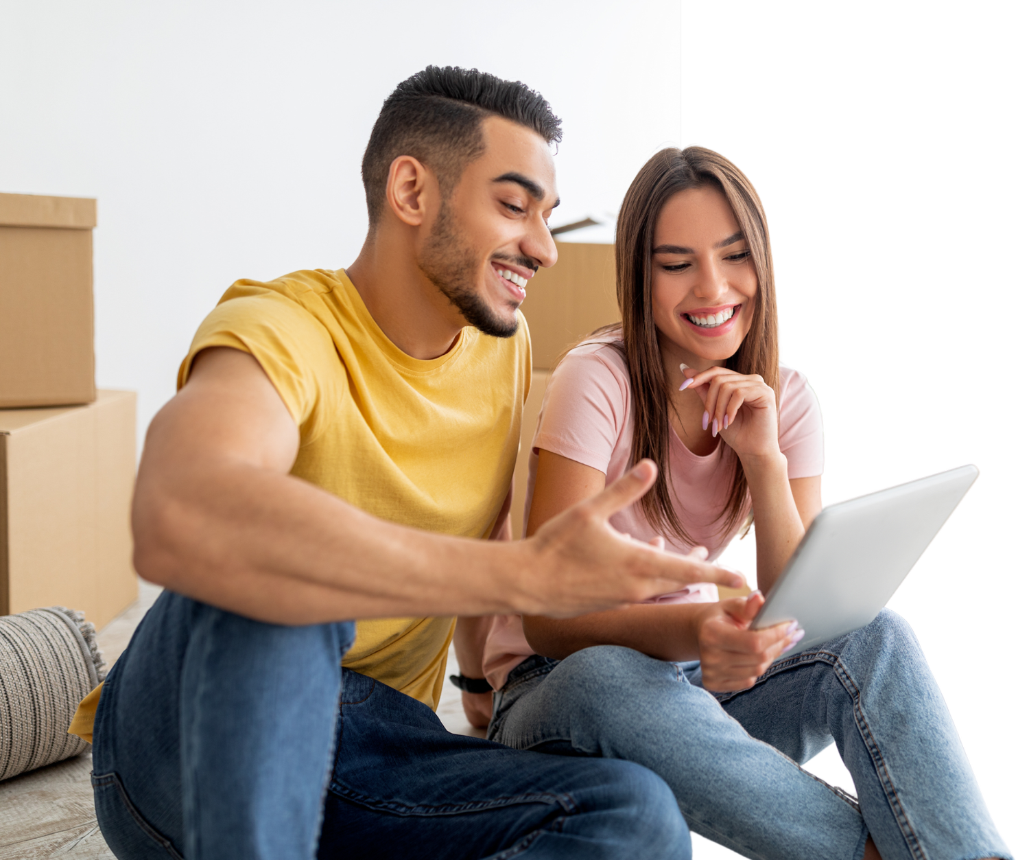 A young couple sitting among moving boxes, smiling as they look at a laptop together