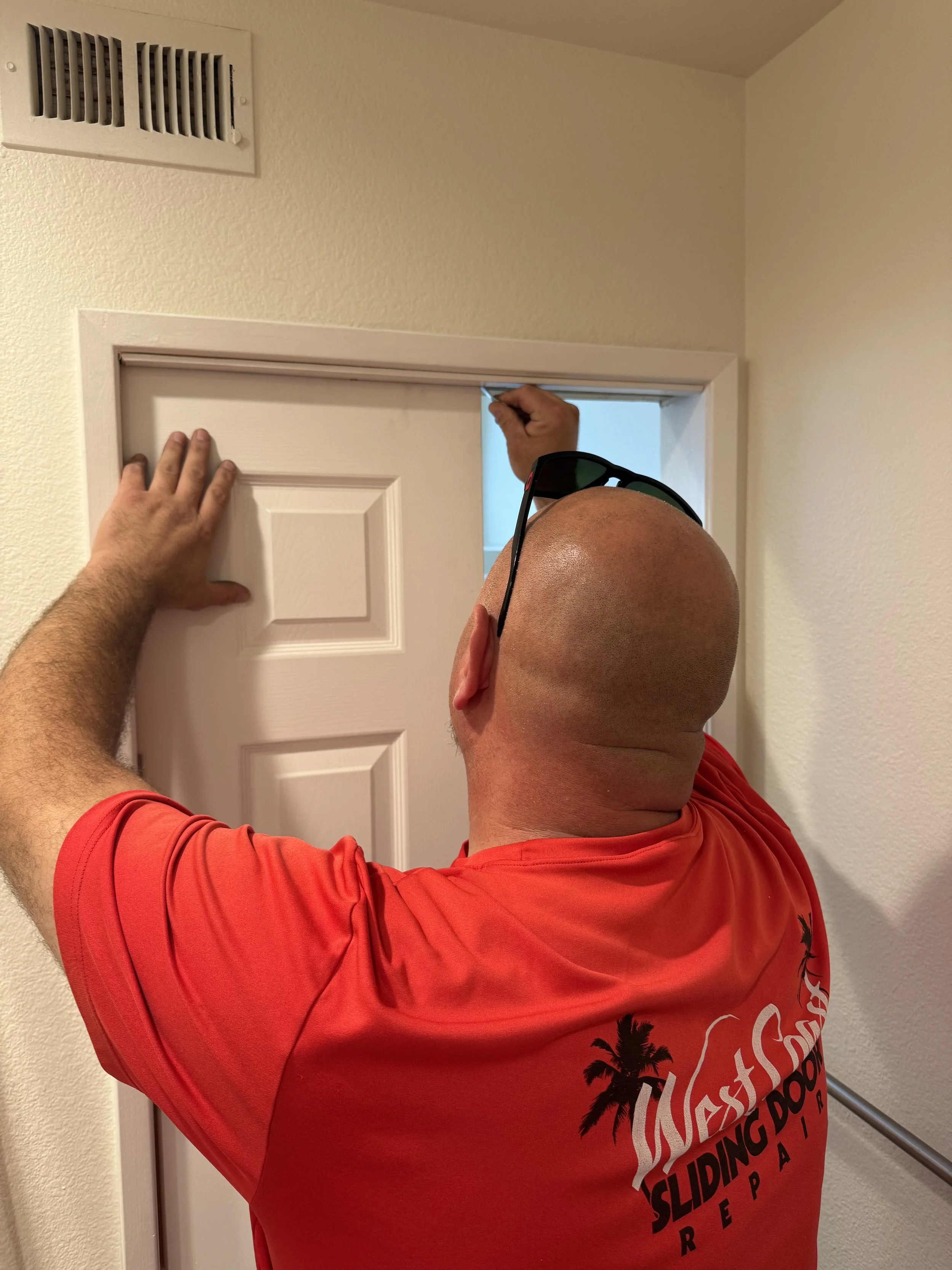 A sliding door repair man in a red t-shirt with palm trees graphic is installing or fixing a door frame while standing on a surface.