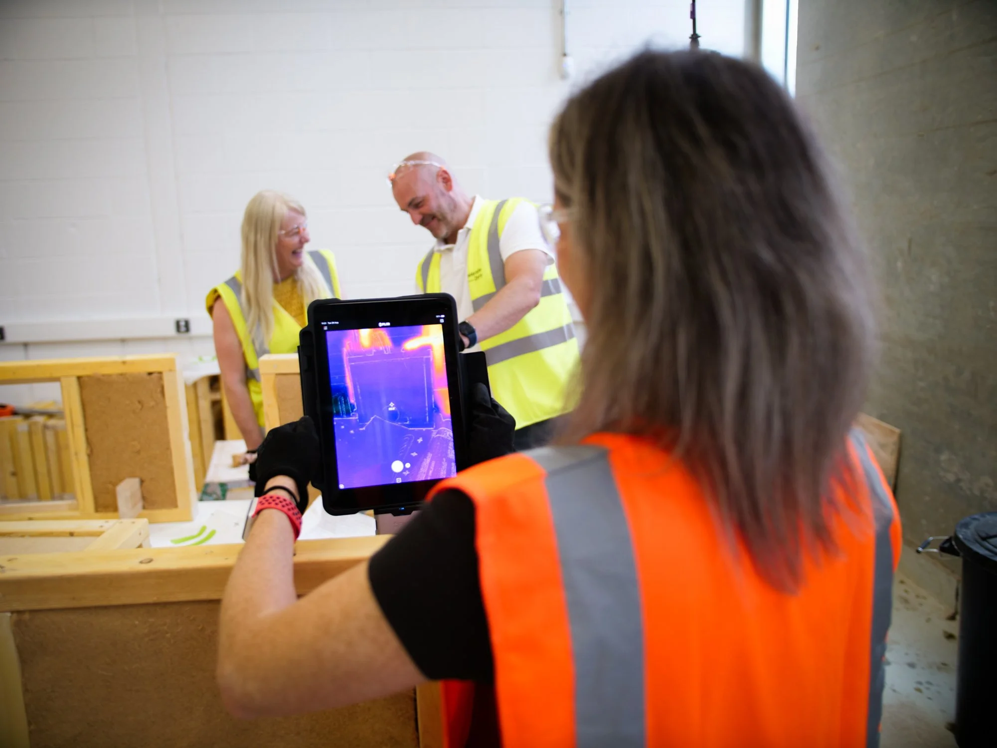 Women in high vis vest, using device to monitor heat installation in wood. A women and man smiling in the background.