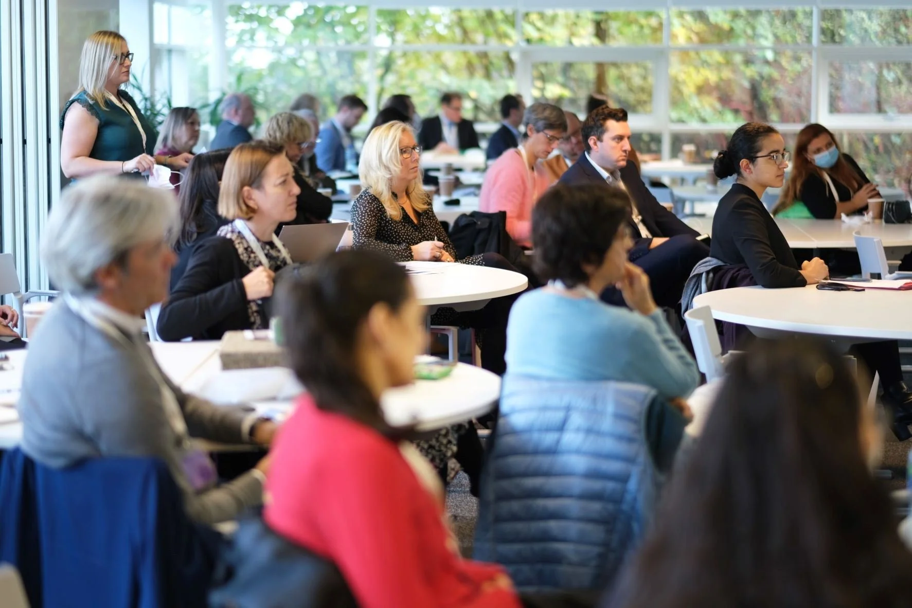 A group of diverse professionals attending a conference or meeting in a bright room with large windows. Some are sitting at tables, others standing, listening attentively.