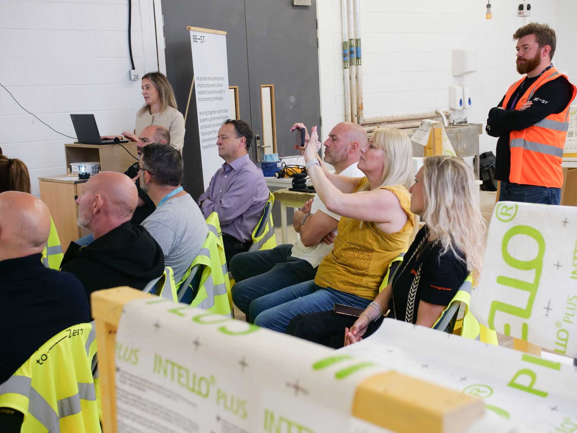 Group of men and women sitting looking at a screen, taking part in a workshop. One women in a yellow top is taking a photo using her phone.