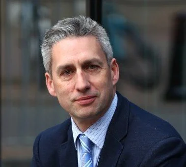 Headshot of a man with gray hair in a suit and tie, outdoors in an urban setting.