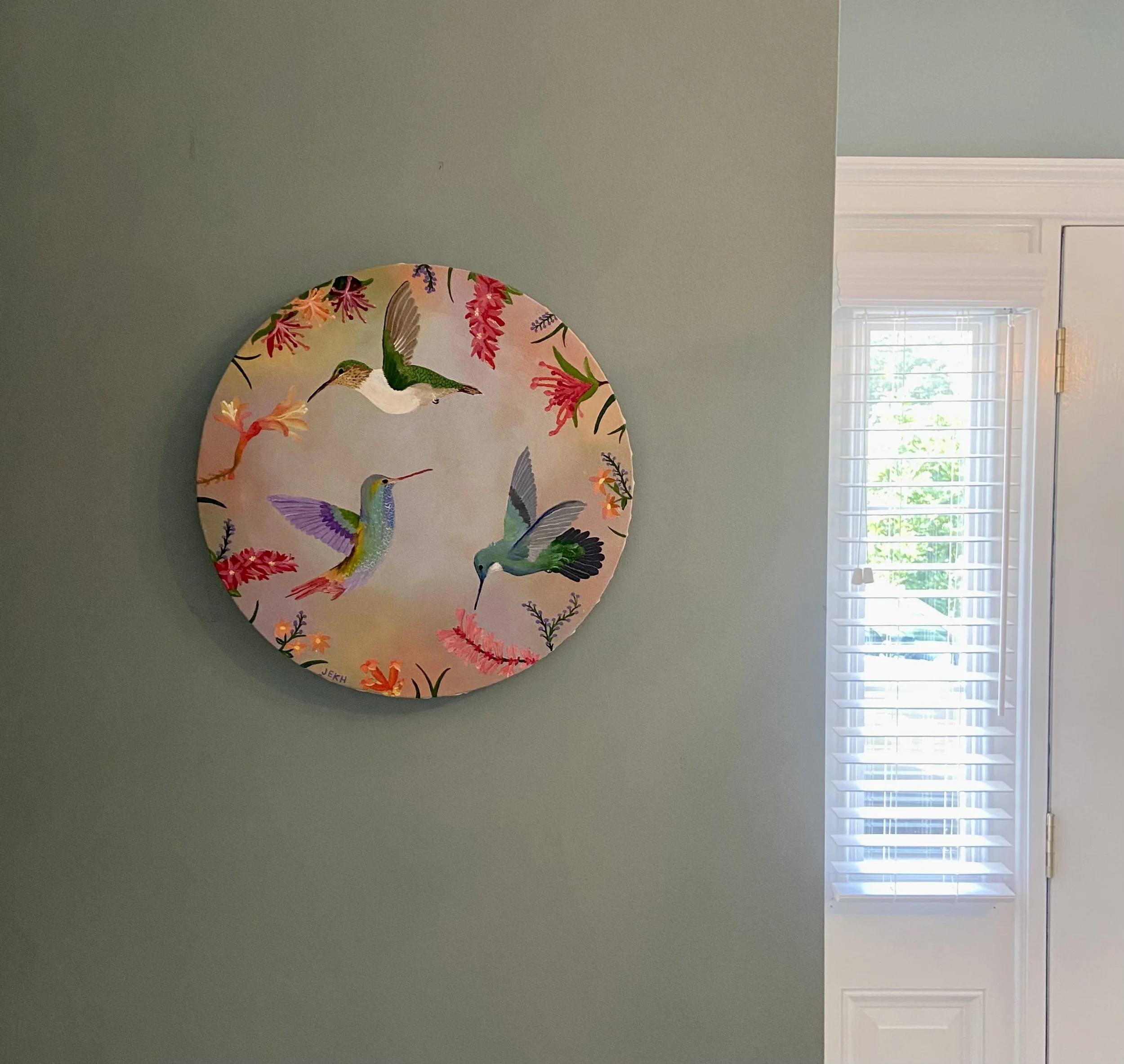 Decorative plate featuring hummingbirds and flowers hanging on a wall near a window with blinds.