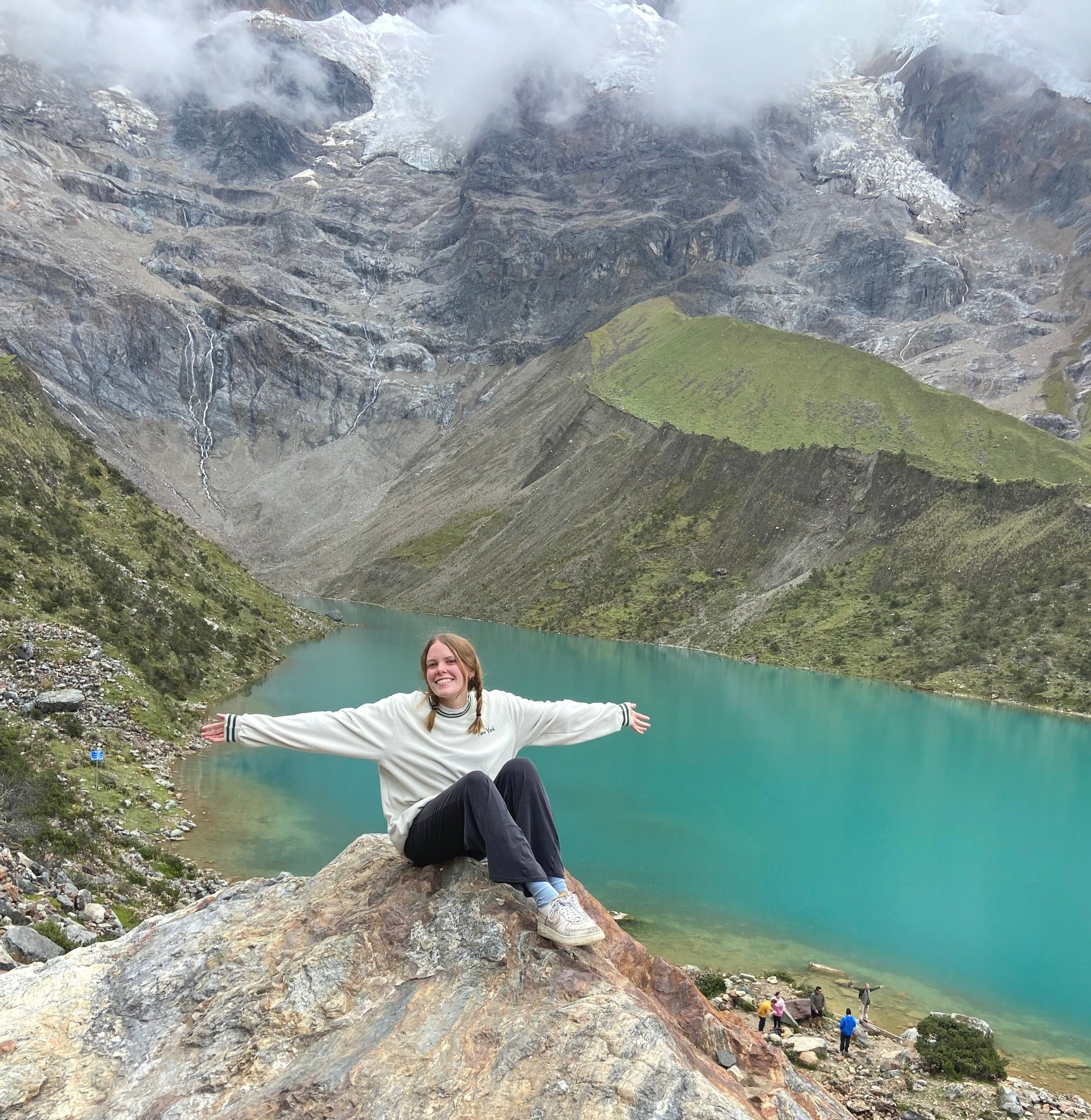 A young woman with braided hair wearing a white sweatshirt and black pants, sitting on a large rock with arms outstretched, smiling in front of a turquoise lake surrounded by mountains with greenery and patches of snow.