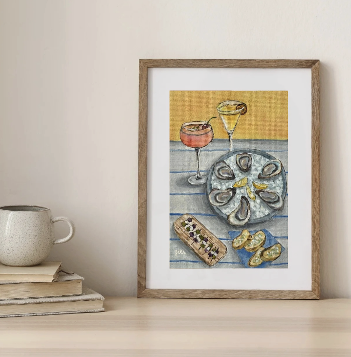 Framed watercolor painting of oysters on a blue-striped surface with two cocktails and a dessert. To the left, a speckled white mug and a stack of books are on a light-colored table.
