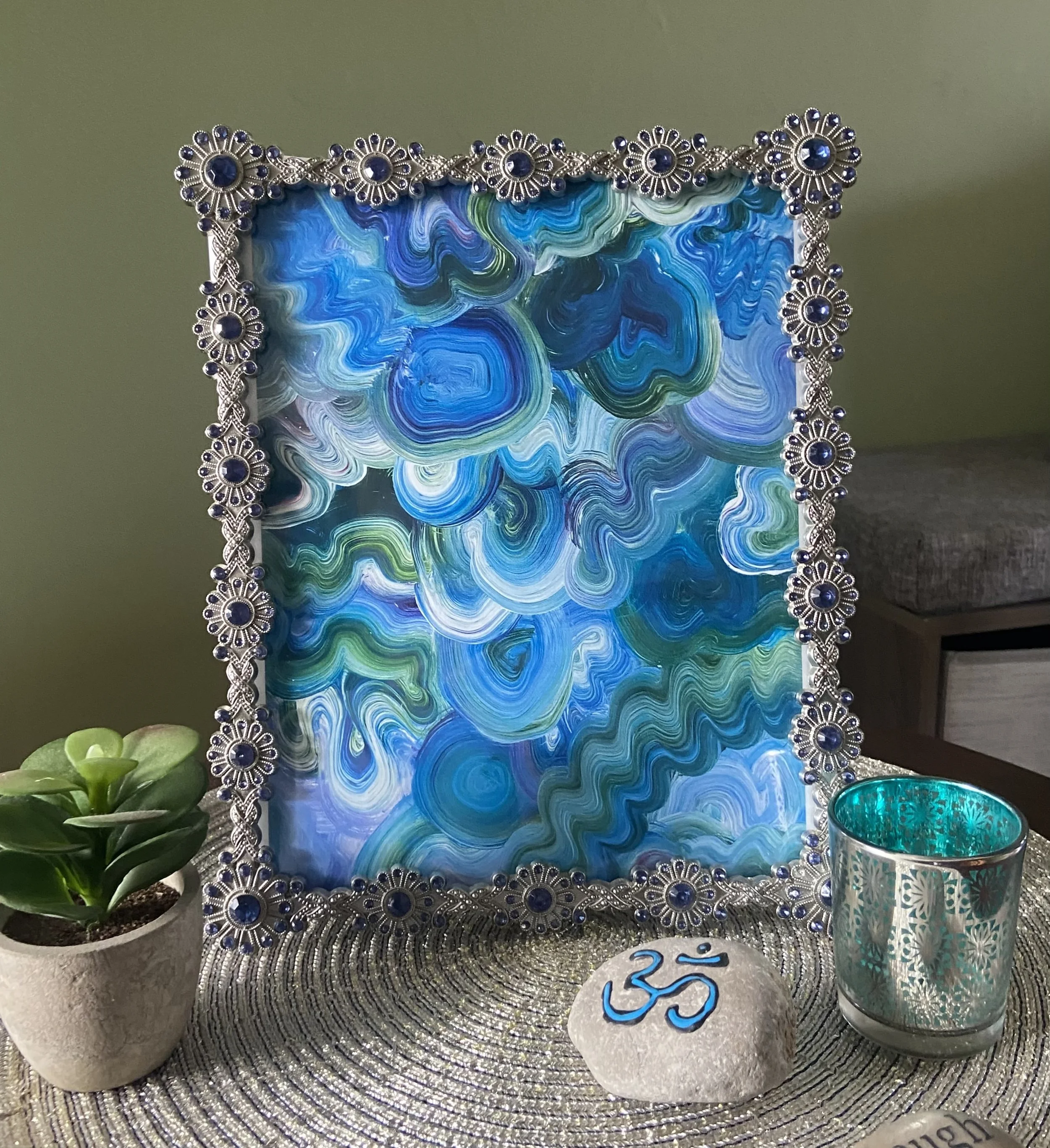 Decorative mirror with a swirling blue and green pattern, surrounded by a silver floral frame with blue gemstones, placed on a textured silver mat. To the left, a green leafy plant in a stone pot, and to the right, a glass containing a turquoise and white patterned candle, with a white stone marker with blue accent markers reading ''LOVE''.