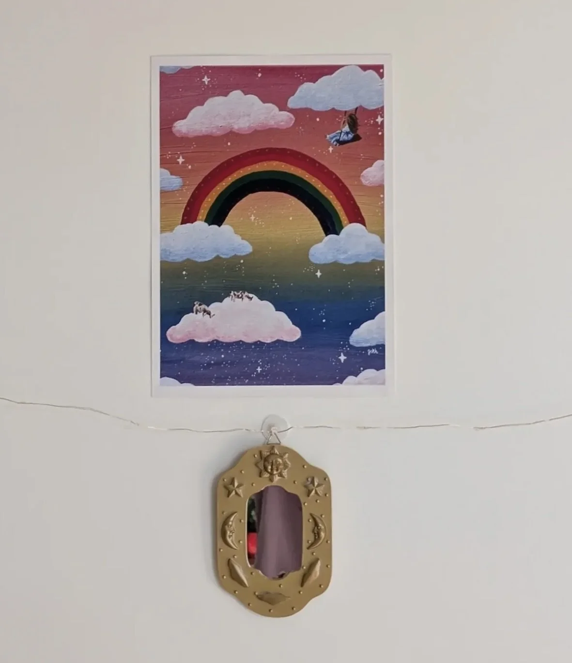 A colorful mural of a rainbow, clouds, and stars with a girl on a swing hanging from a cloud, above clouds with zebras and stars in a gradient sky. Below, a small mirror hangs on a white wall.