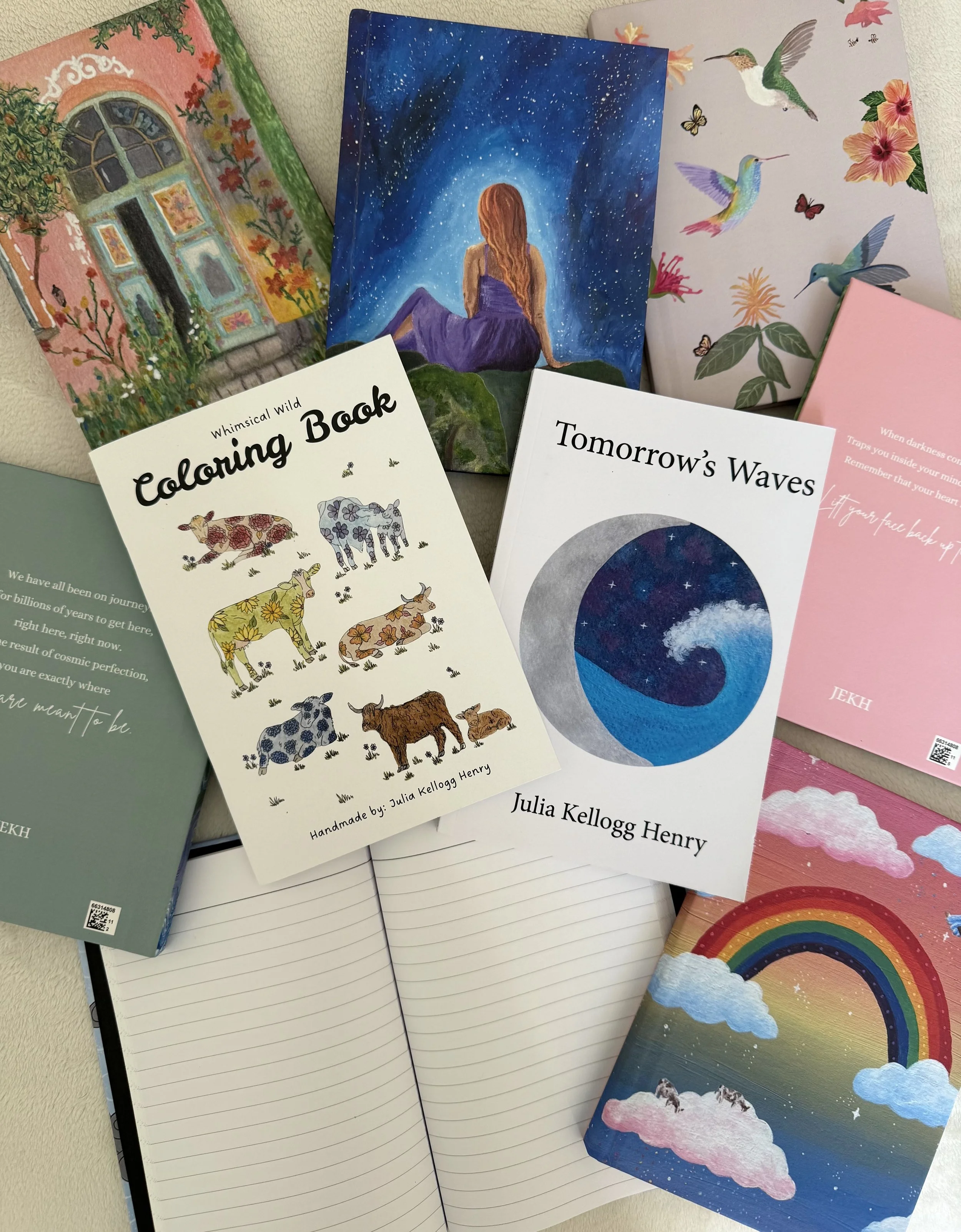 Collection of colorful notebooks and journals featuring artistic covers, including a girl sitting under a starry sky, hummingbirds with vibrant flowers and butterflies, a rainbow and cloud scene, and illustrations of cows with floral patterns. Some books have printed titles, such as 'Coloring Book' and 'Tomorrow's Waves,' along with author names Julia Kellogg Henry and JEKH.