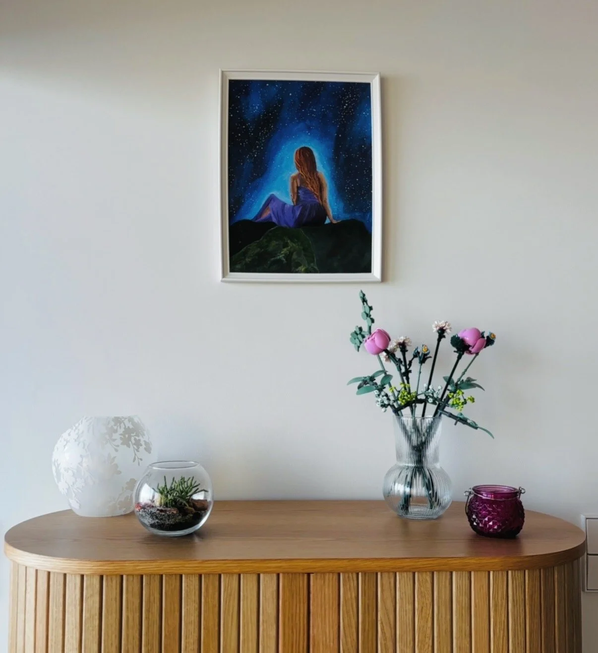 A wooden sideboard with a floral arrangement in a glass vase, a pink candle holder, and a white decorative lamp with a floral pattern. A framed painting of a woman with long hair sitting on a rock under a starry night sky hangs on the white wall above.
