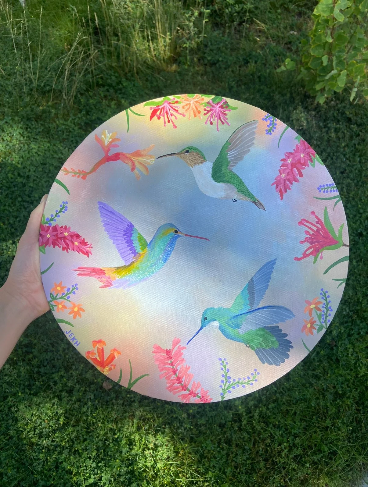A round decorative plate with colorful illustrations of four hummingbirds surrounded by vibrant pink, purple, orange, and blue flowers, held outdoors over green grass.