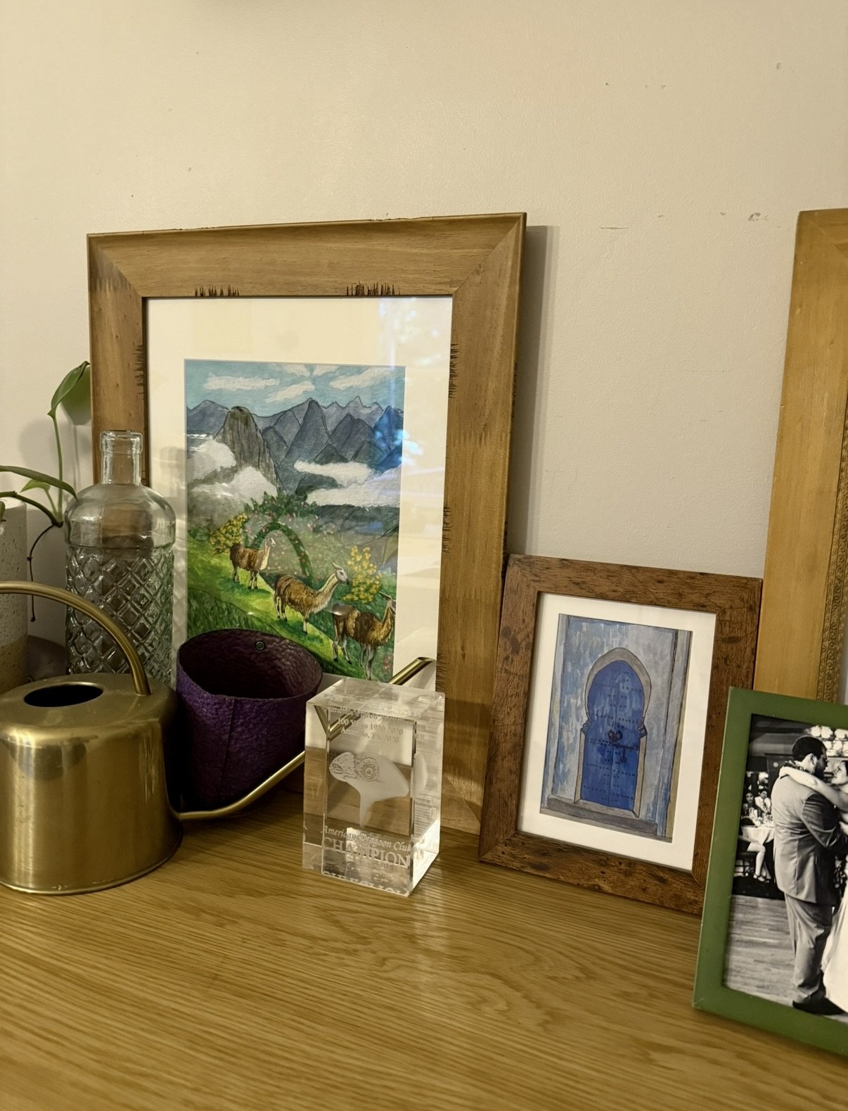 Decorative tabletop with framed pictures, a glass jar, a purple candle holder, and a gold watering can, against a plain wall.