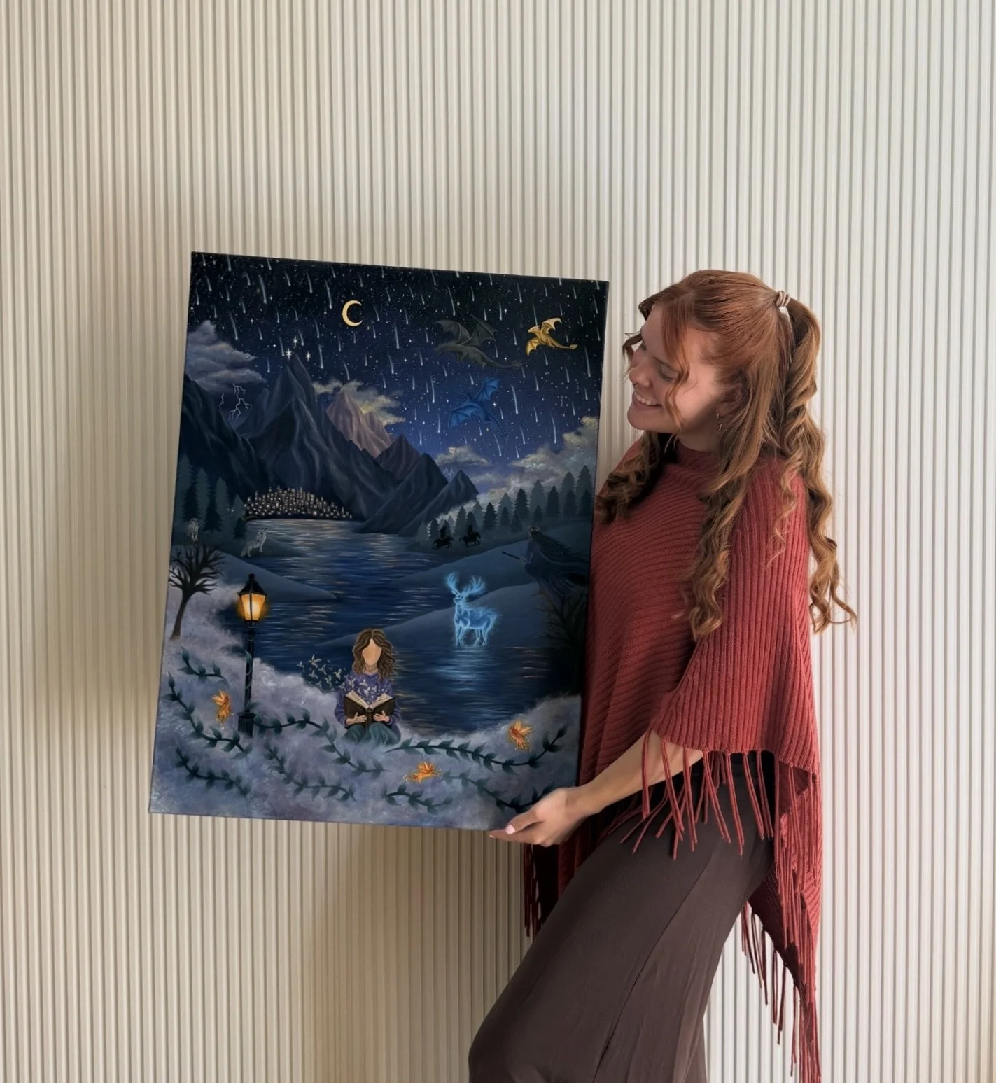 A young woman with long red hair, wearing a red fringed shawl and a brown skirt, smiling while holding a large fantasy-themed painting. The painting depicts a mountainous landscape under a starry night sky with falling stars, a crescent moon, dragons, a glowing blue deer, a girl reading a book beside a lantern, and a river reflecting the starry sky.