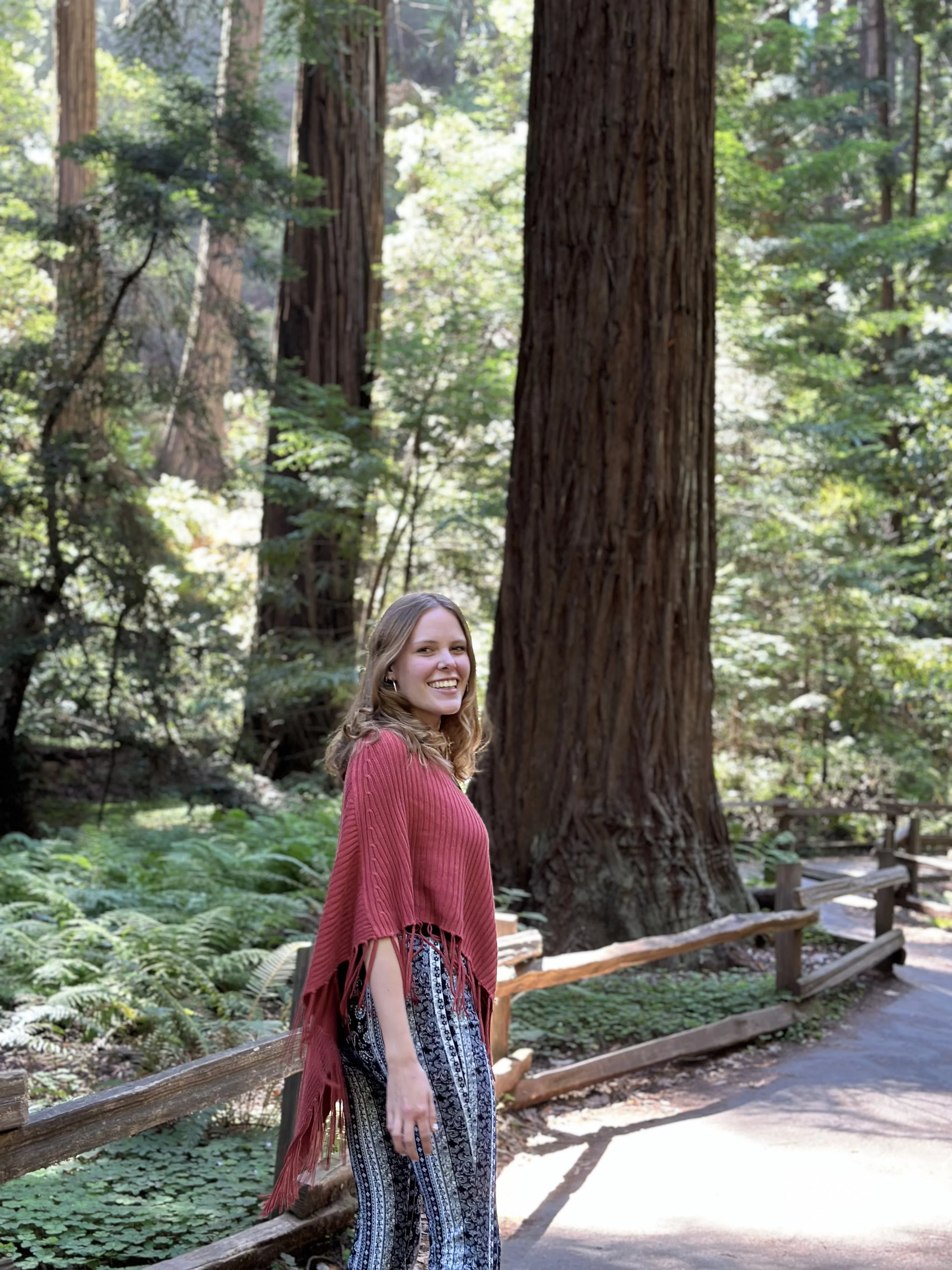 A woman with light brown hair smiling in a red poncho and patterned pants standing on a forest path surrounded by tall redwood trees and green foliage.