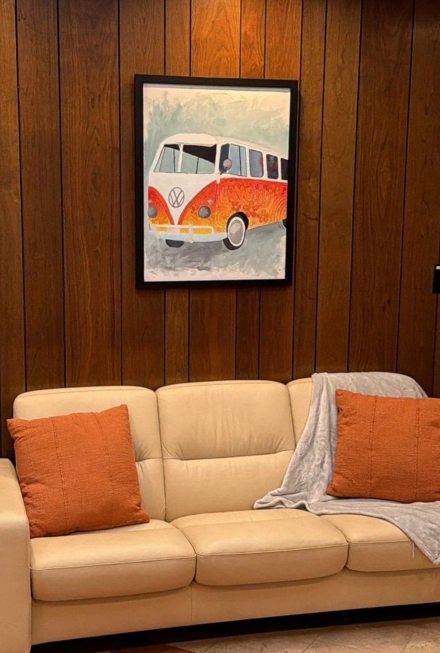 A beige leather couch with two orange pillows and a gray blanket on it, set against a wooden paneled wall with a framed painting of a vintage Volkswagen bus in front of the wall.