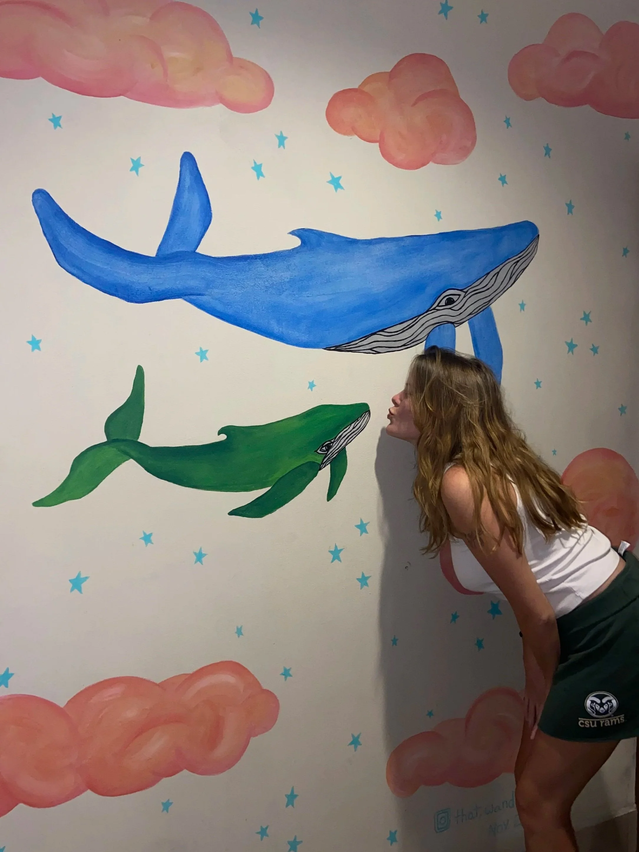 A colorful wall mural of whales swimming among pink clouds and blue stars. A person with long hair is leaning against the wall, partially blocking one whale.