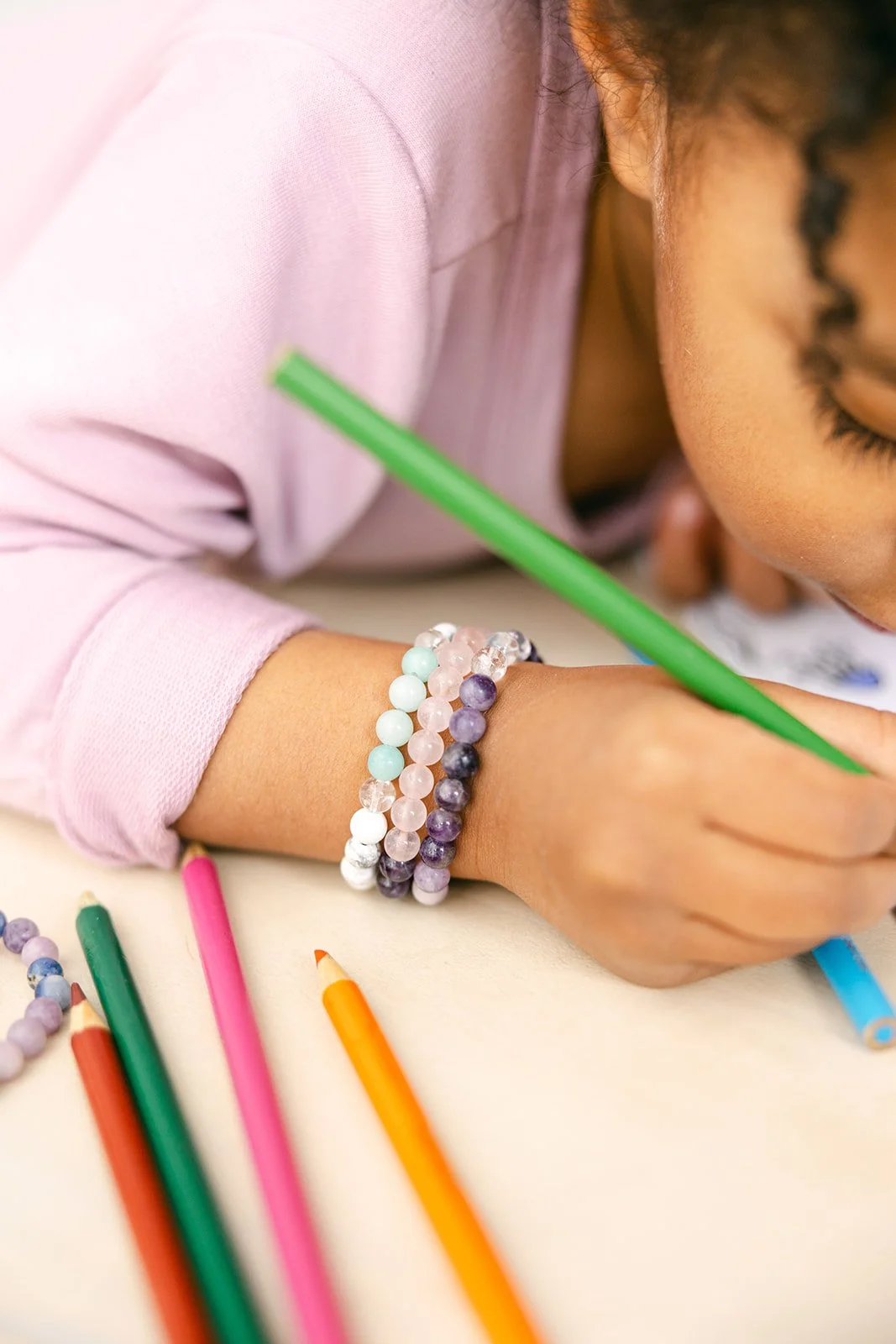A child coloring with green crayon, wearing pastel-colored beaded bracelets, with colored pencils scattered on the table.
