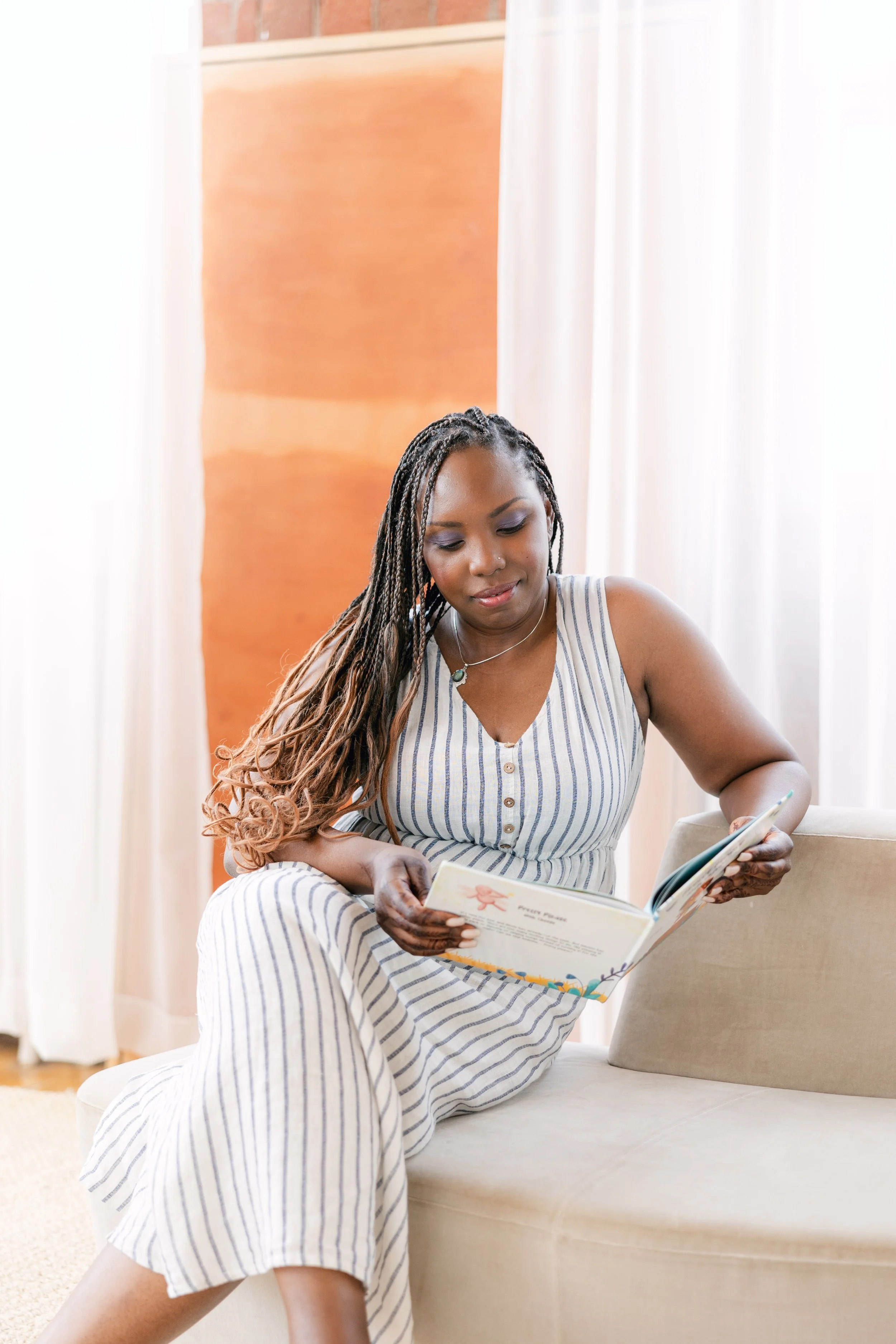 A woman with long braids and a striped sleeveless dress reading a book while sitting on a beige couch in a bright room with white curtains.