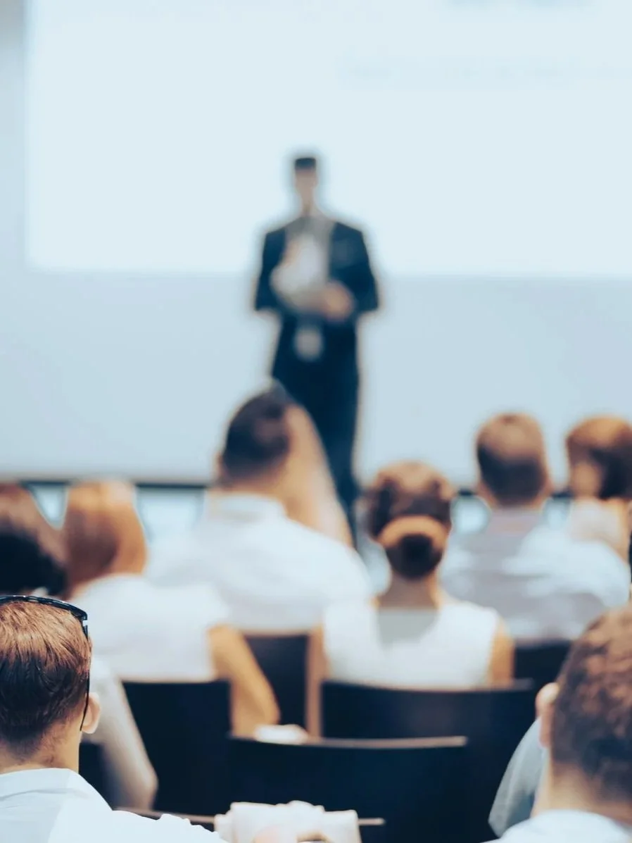 A blurry image of a person standing in front of an audience, giving a presentation or speech in a conference room.