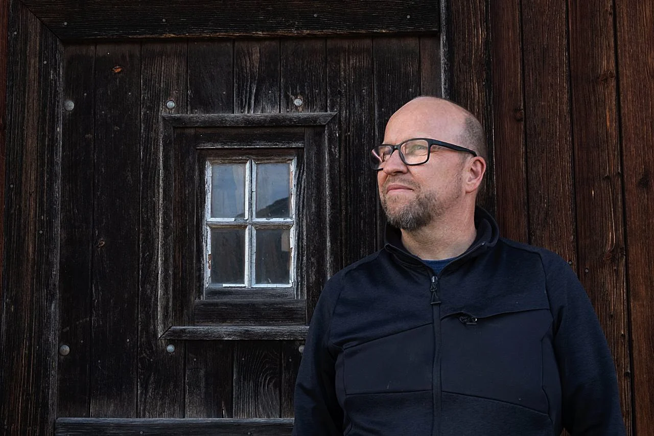 Un homme à lunettes et barbe, portant une veste noire, se tient devant une façade en bois sombre avec une petite fenêtre fixture.