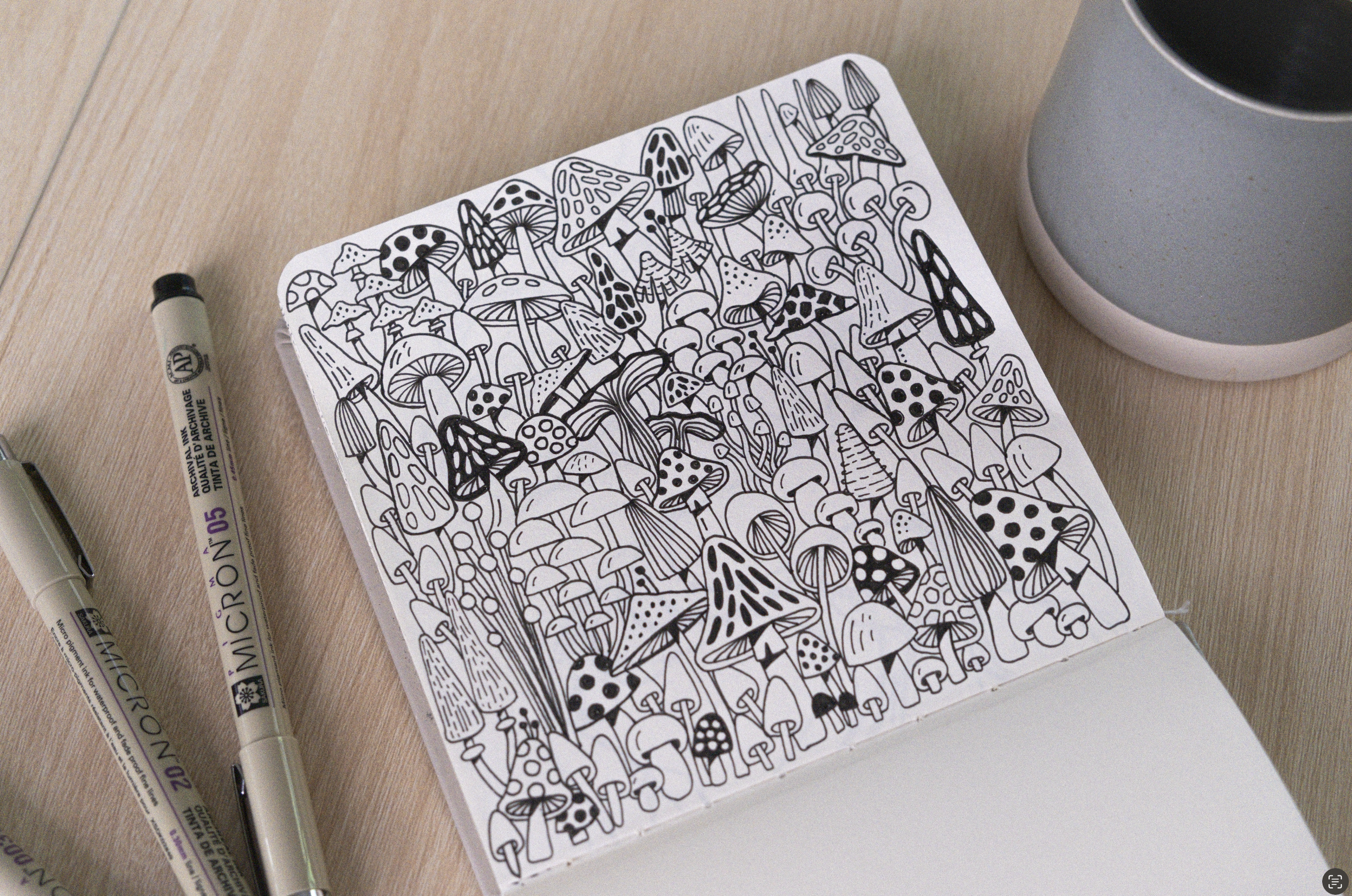 A black and white coloring page featuring various mushrooms. The page is lying on a wooden surface, with two black markers beside it and a gray cup partially visible in the top right corner.