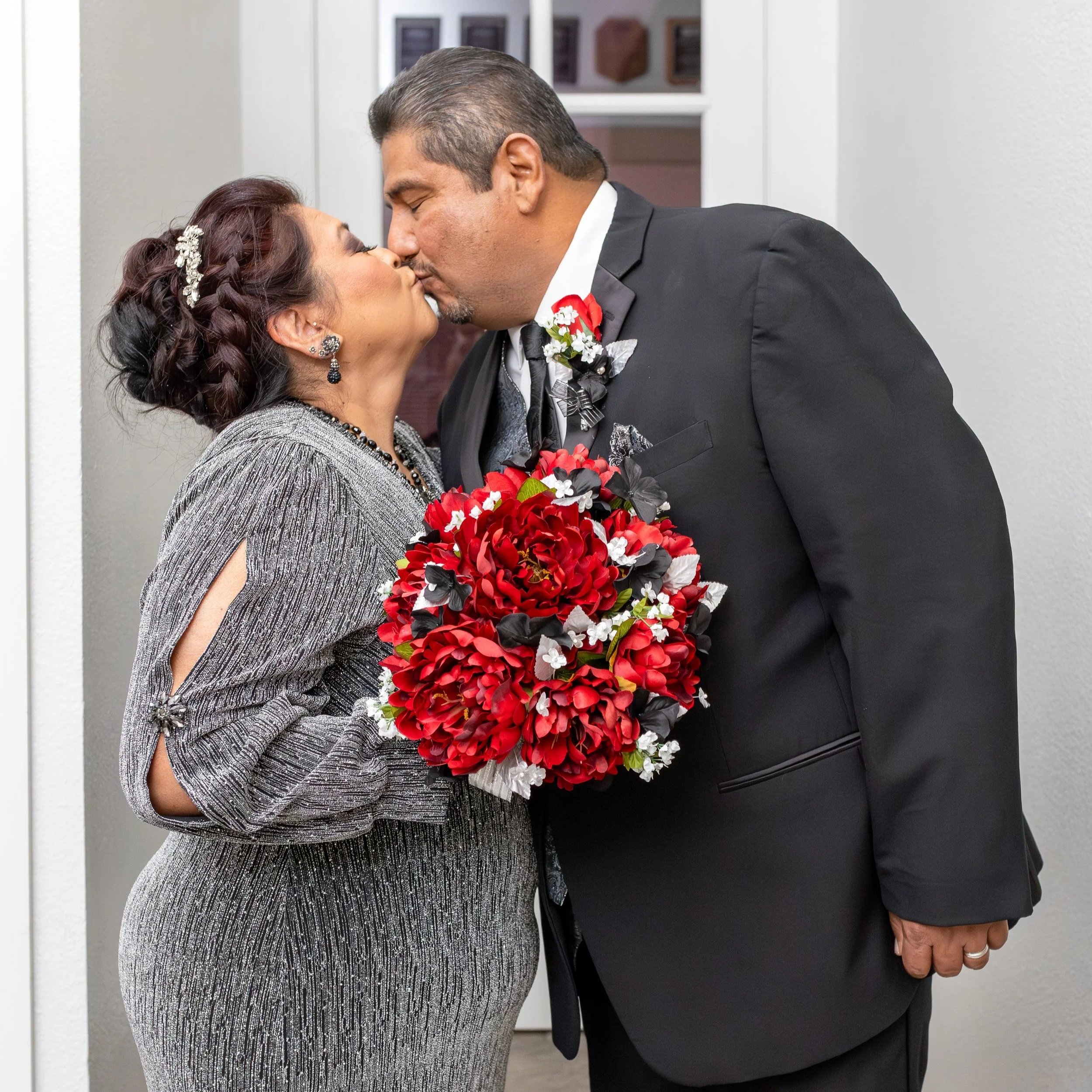 A couple dressed in formal attire sharing a kiss, with the woman holding a bouquet of red and white flowers.
