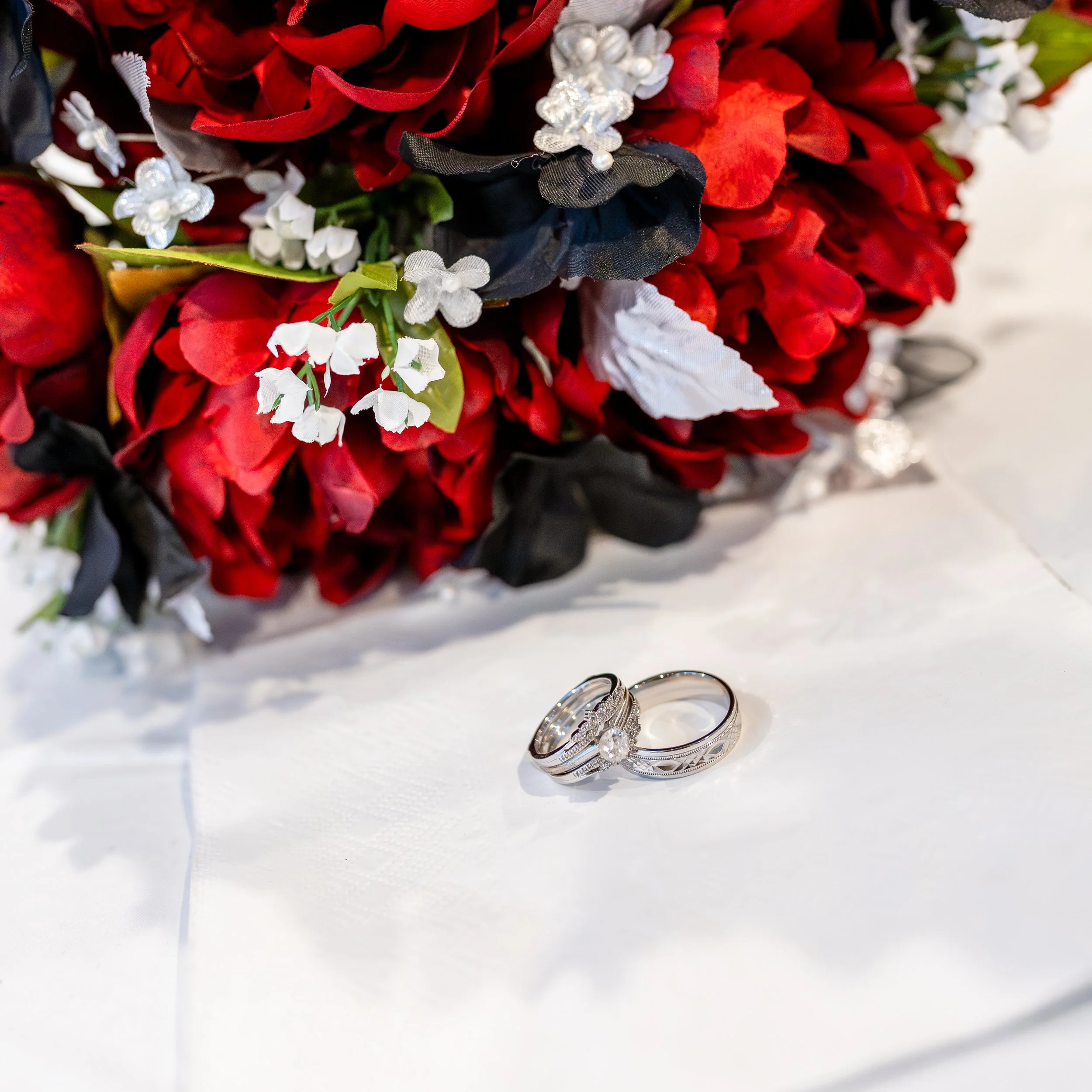 Silver wedding rings placed on a white tablecloth in front of a large bouquet of red, black, and white flowers.