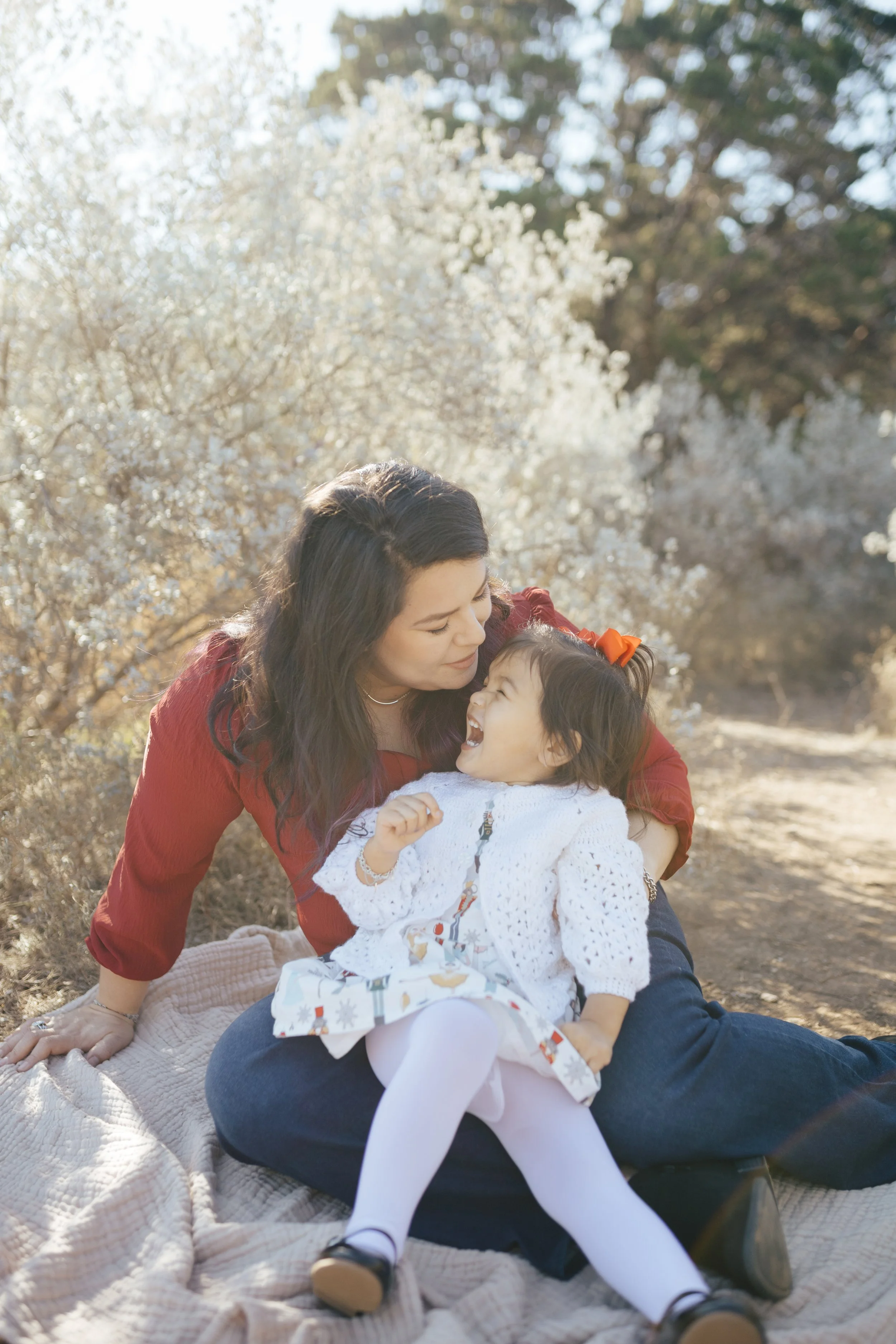 A woman with dark hair and a young girl with a bow in her hair sharing a joyful moment outdoors on a blanket, surrounded by blooming trees.