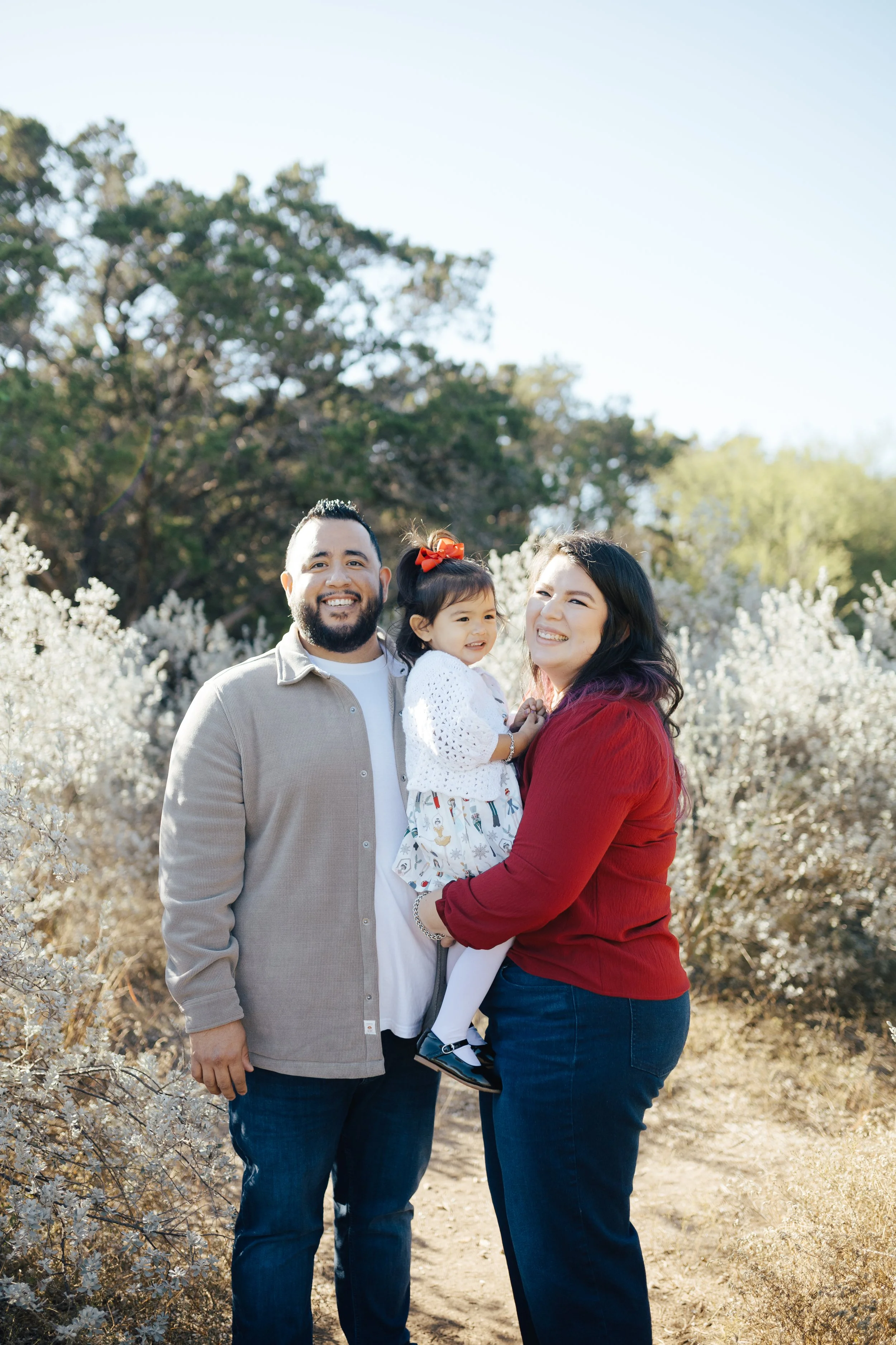 A smiling family of three outdoors with bushes and trees in the background - a man, woman, and young girl.