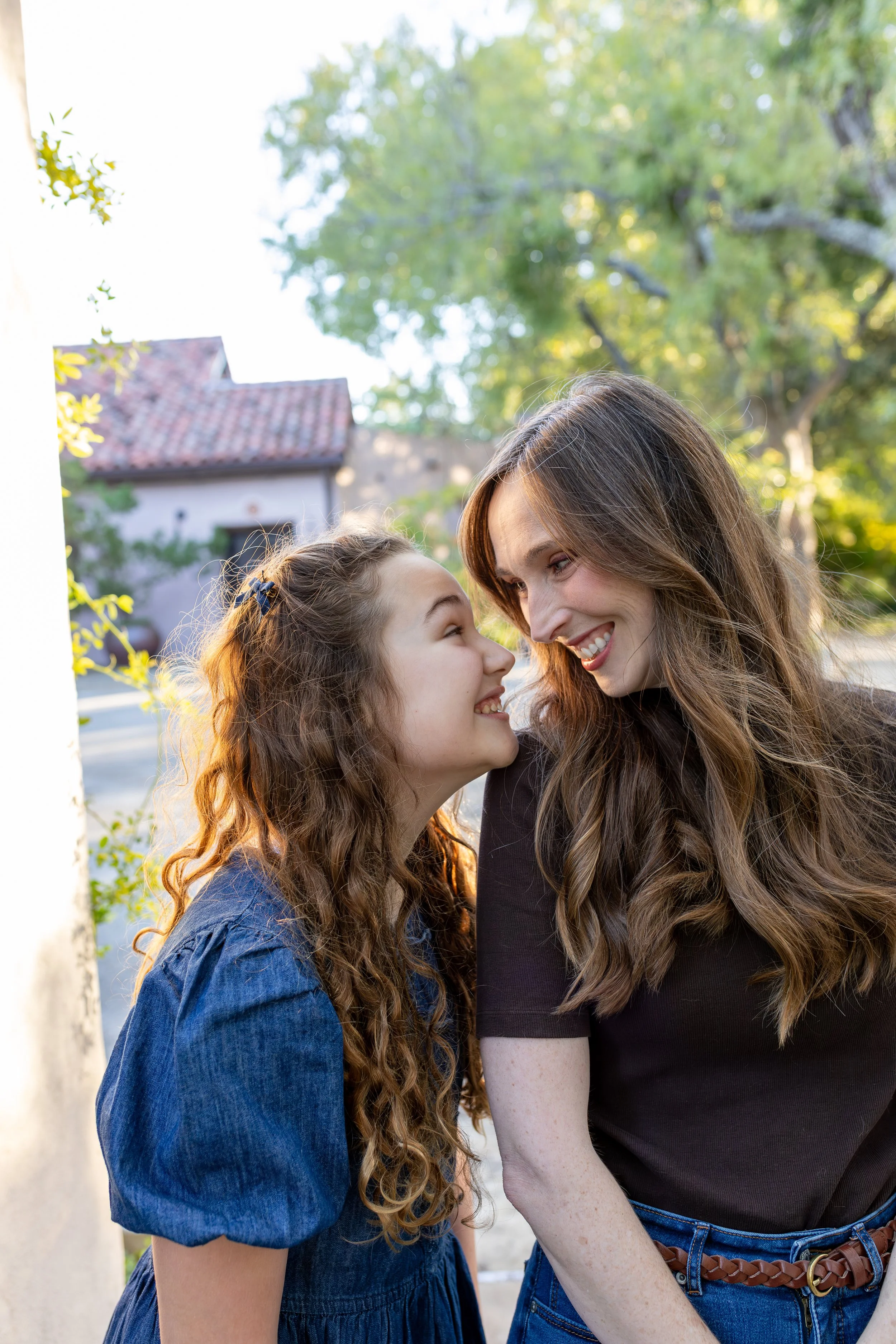 A woman and a girl with long, curly hair smiling at each other outdoors on a sunny day.