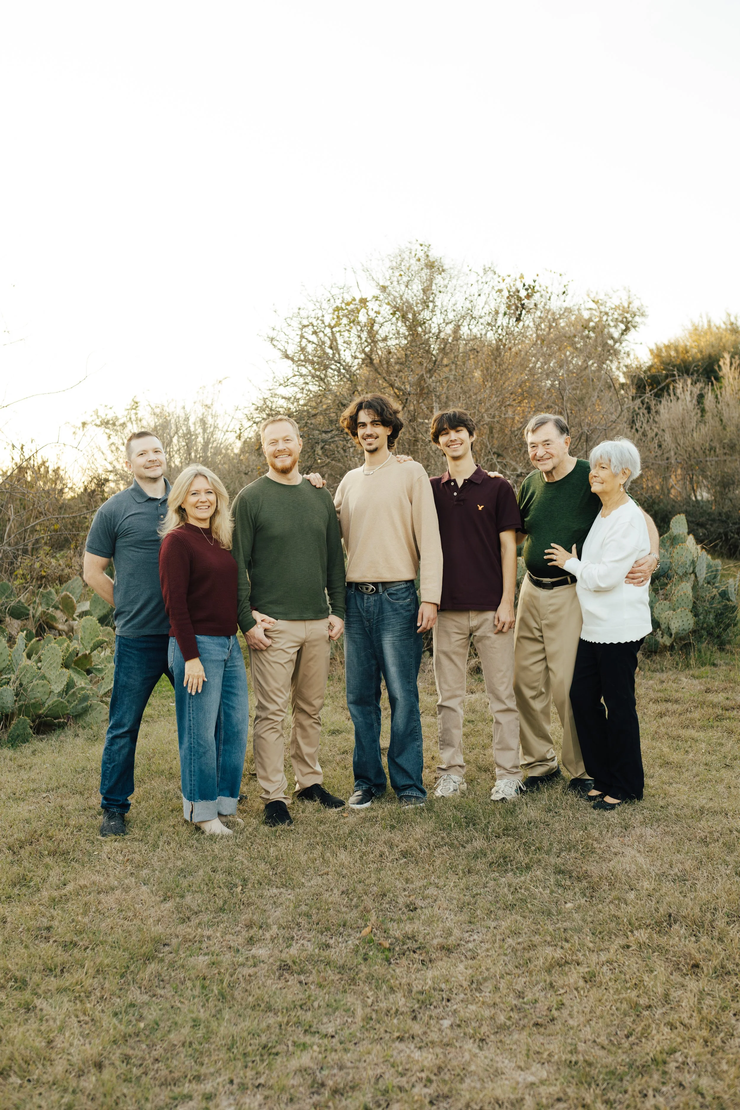 Group of eight people, including three elderly, standing outdoors on grass with trees and cacti in the background, smiling at the camera.