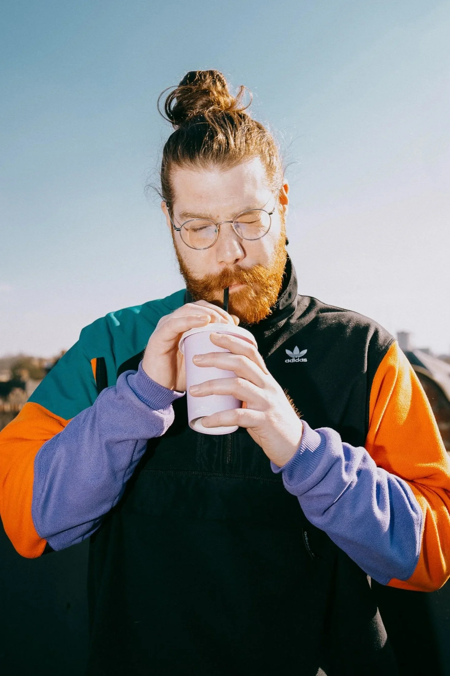A man with glasses and a red beard wearing a colorful jacket drinks from a paper cup with a straw outdoors.