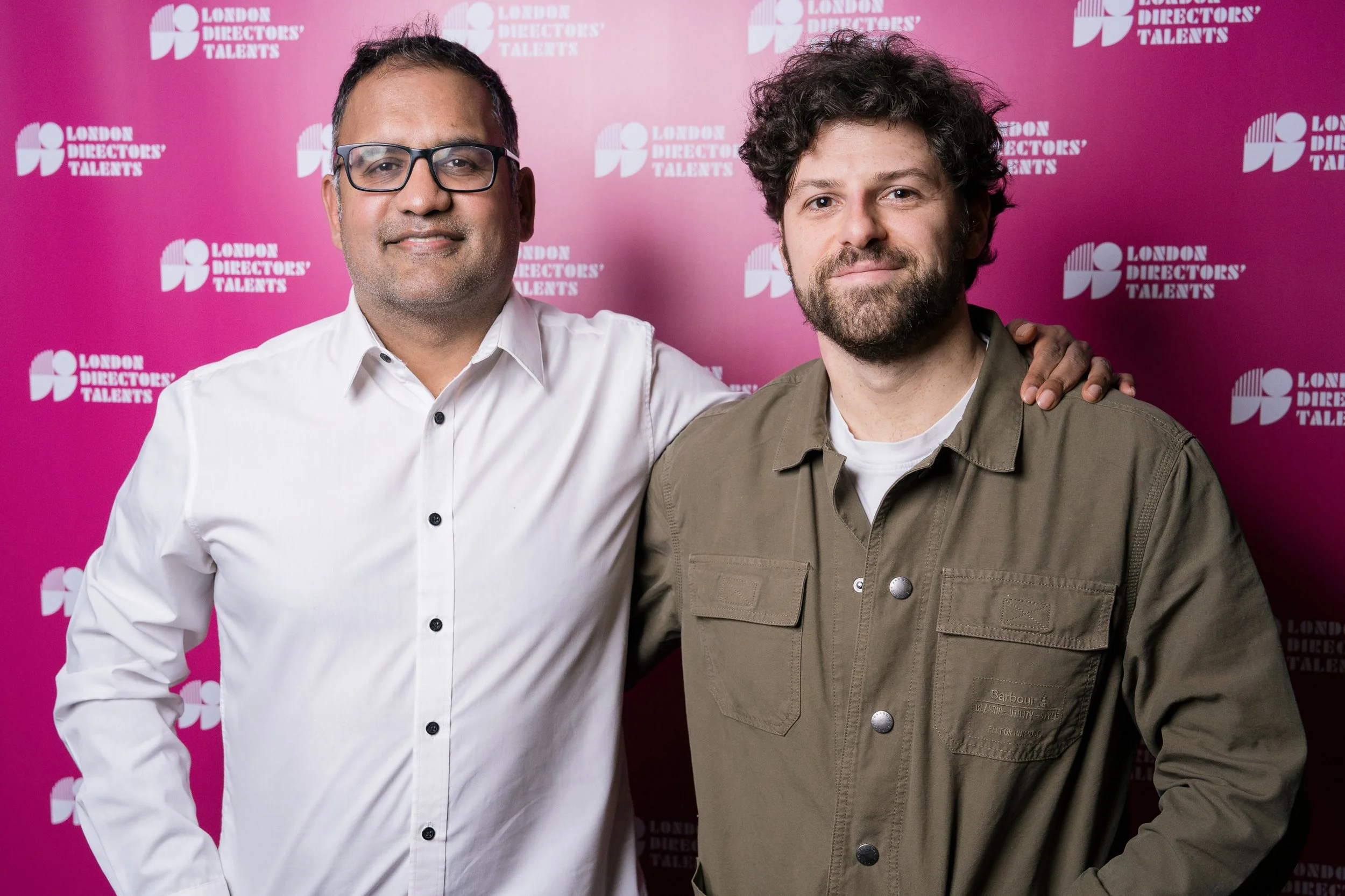Two men standing together in front of a pink backdrop with white text that reads 'London Directors' Talents'. One man is wearing a white shirt and glasses, and the other man is wearing an olive green jacket.