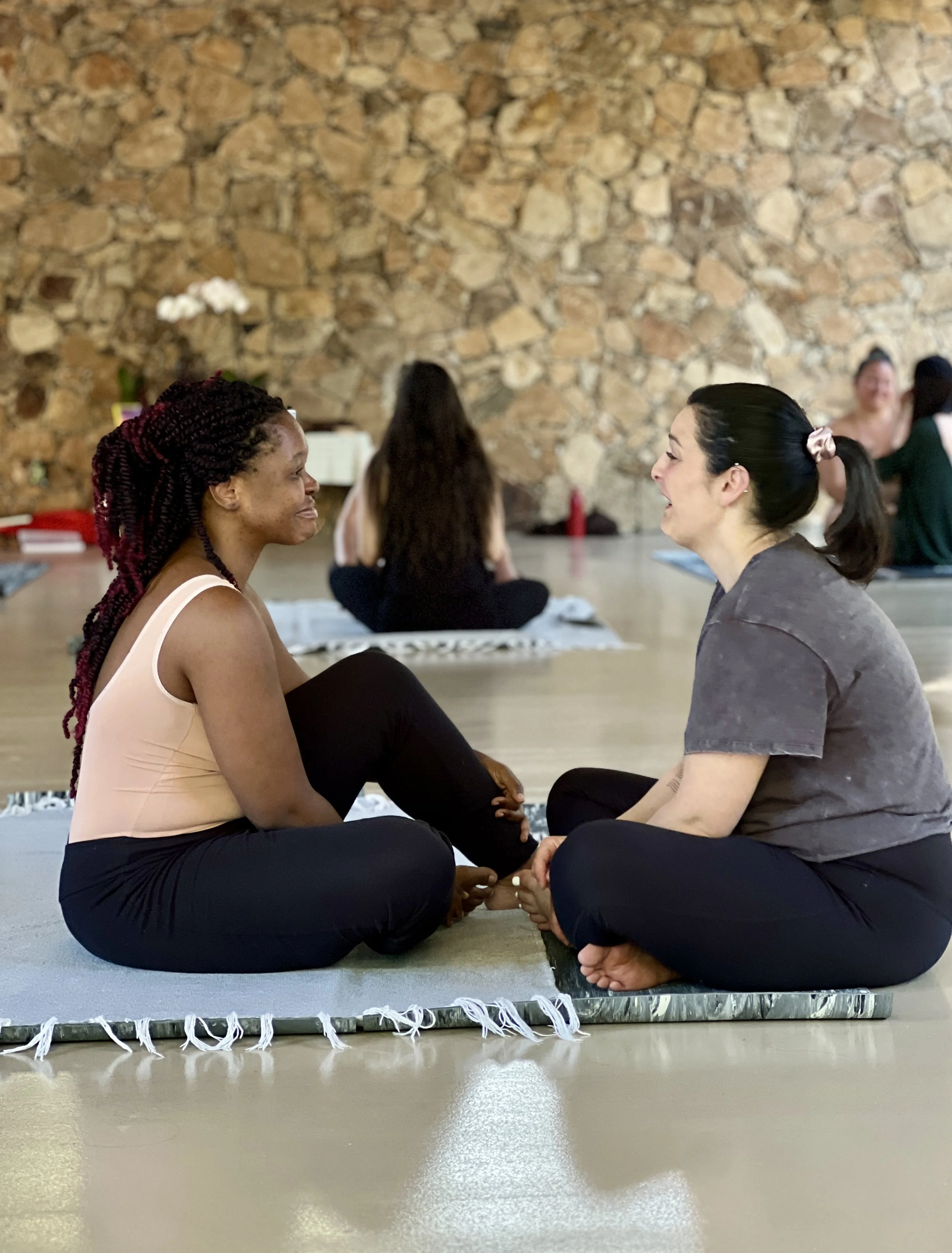Two women sit cross-legged on yoga mats, facing each other and smiling during a yoga or meditation class in a studio with a stone wall background.