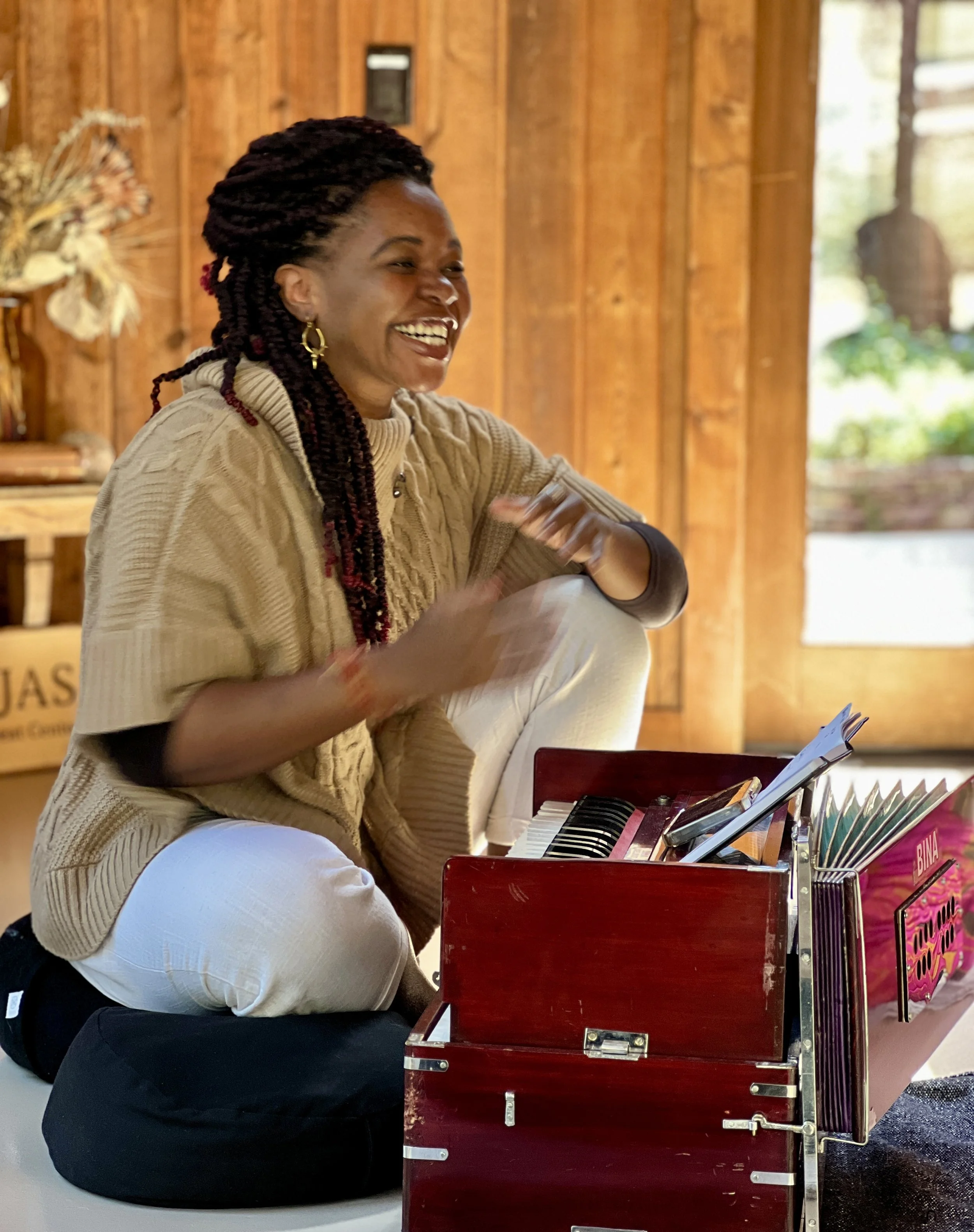 Woman with red and black braided hair, wearing a beige sweater, sitting on a black cushion, laughing, next to a red miniature piano with sheet music, inside a wooden room with a door and window.