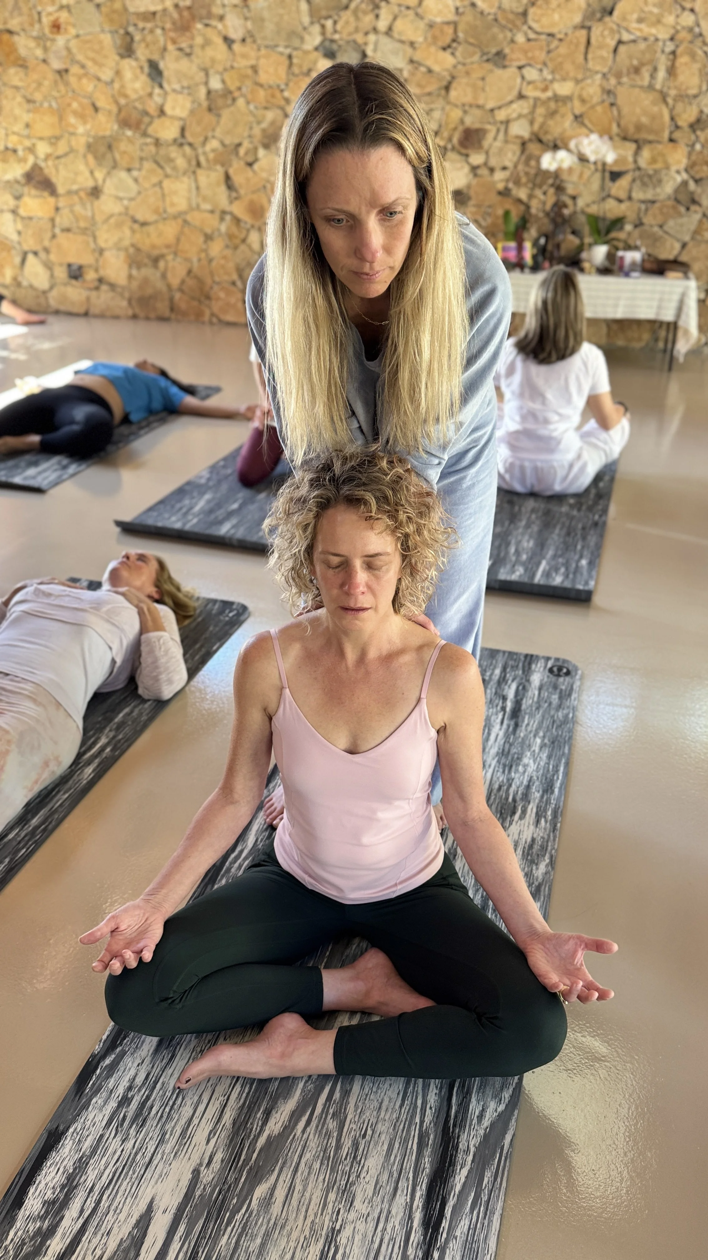 A woman practicing yoga in a meditation pose with a yoga instructor guiding her, in a studio with other participants lying on mats in the background.