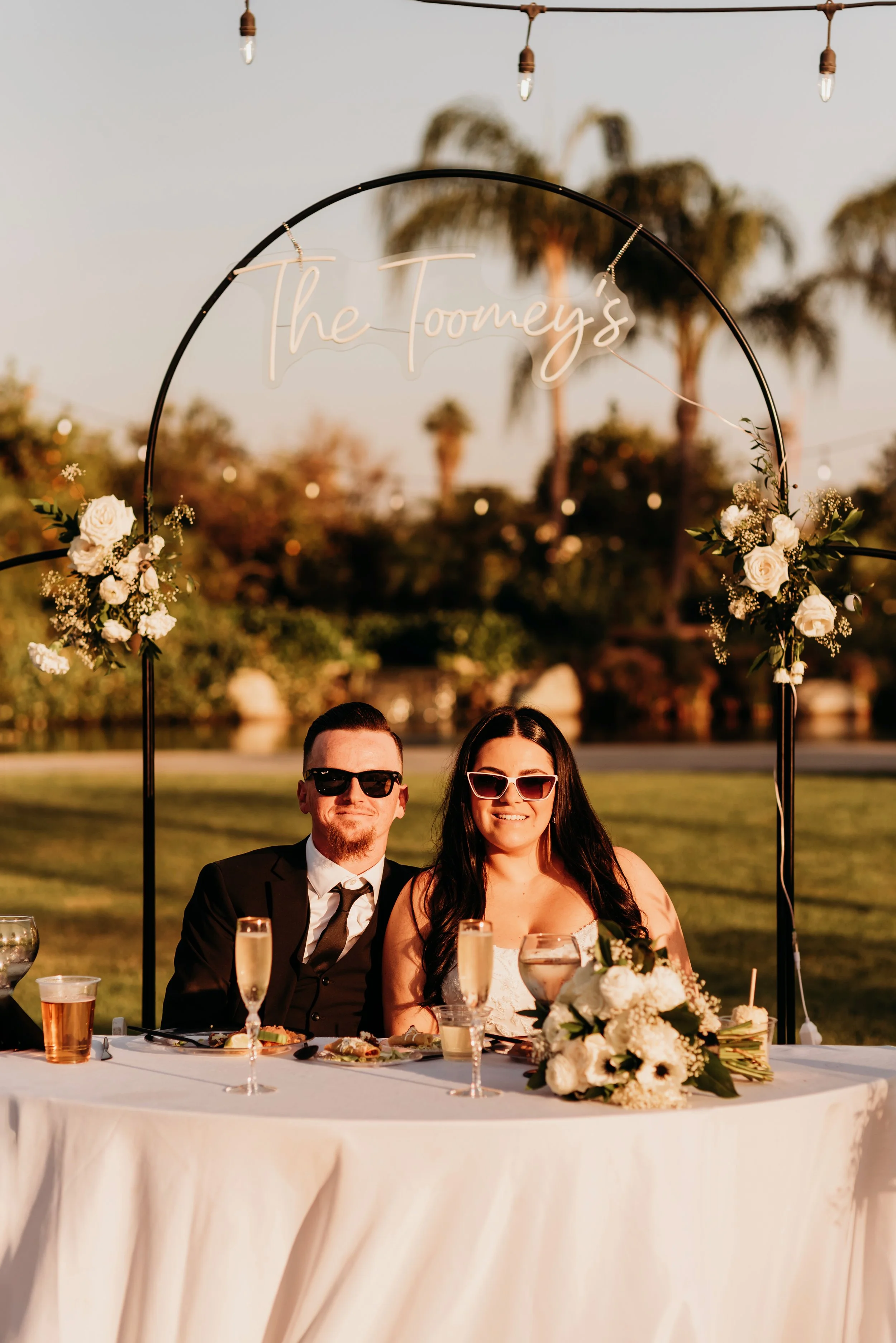 A couple dressed in wedding attire sitting at a decorated reception table outdoors during sunset, with a sign above them reading 'The Tooney's' and string lights overhead.