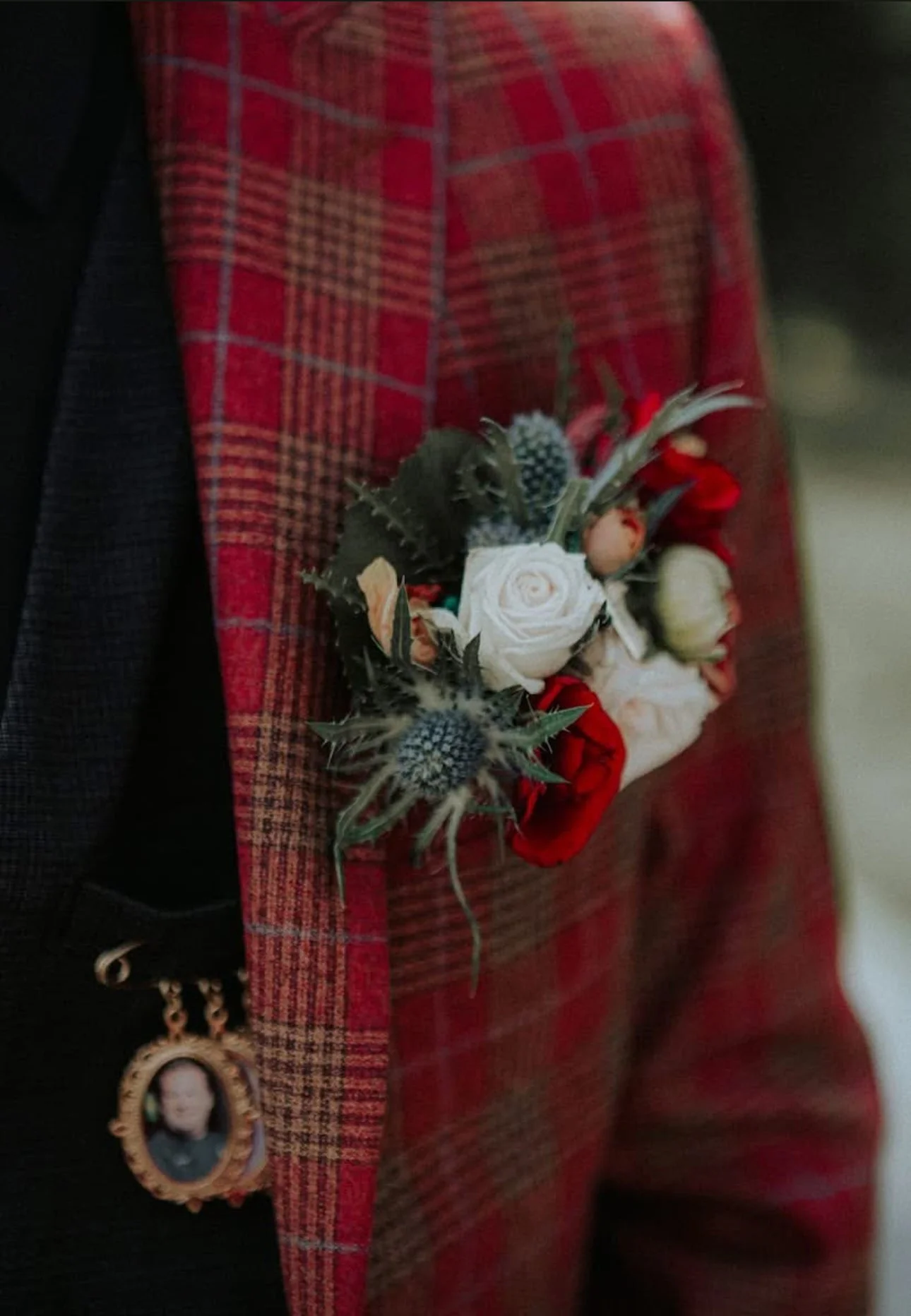 Person wearing a red plaid jacket with a small floral corsage pinned to the lapel, along with a portrait charm hanging from the same lapel.